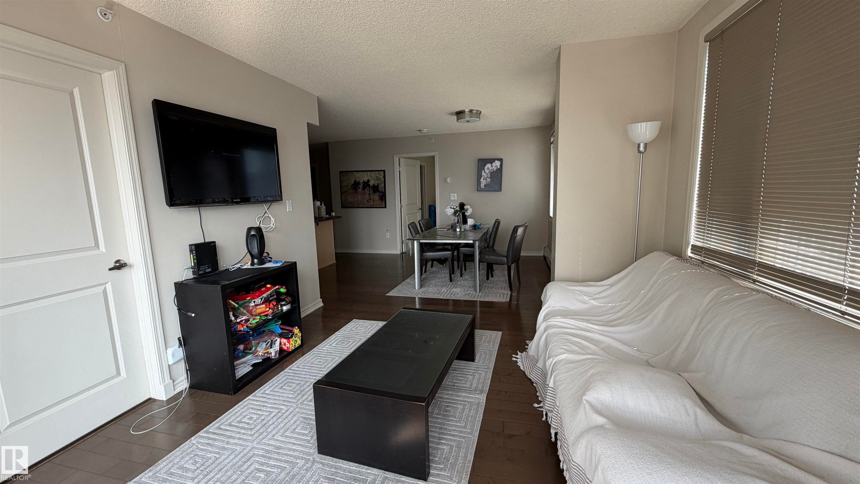 Open-concept living area with wood-finish flooring, neutral wall tones, and horizontal window blinds - #2401 10180 104 St Nw, Edmonton, AB - Indoor