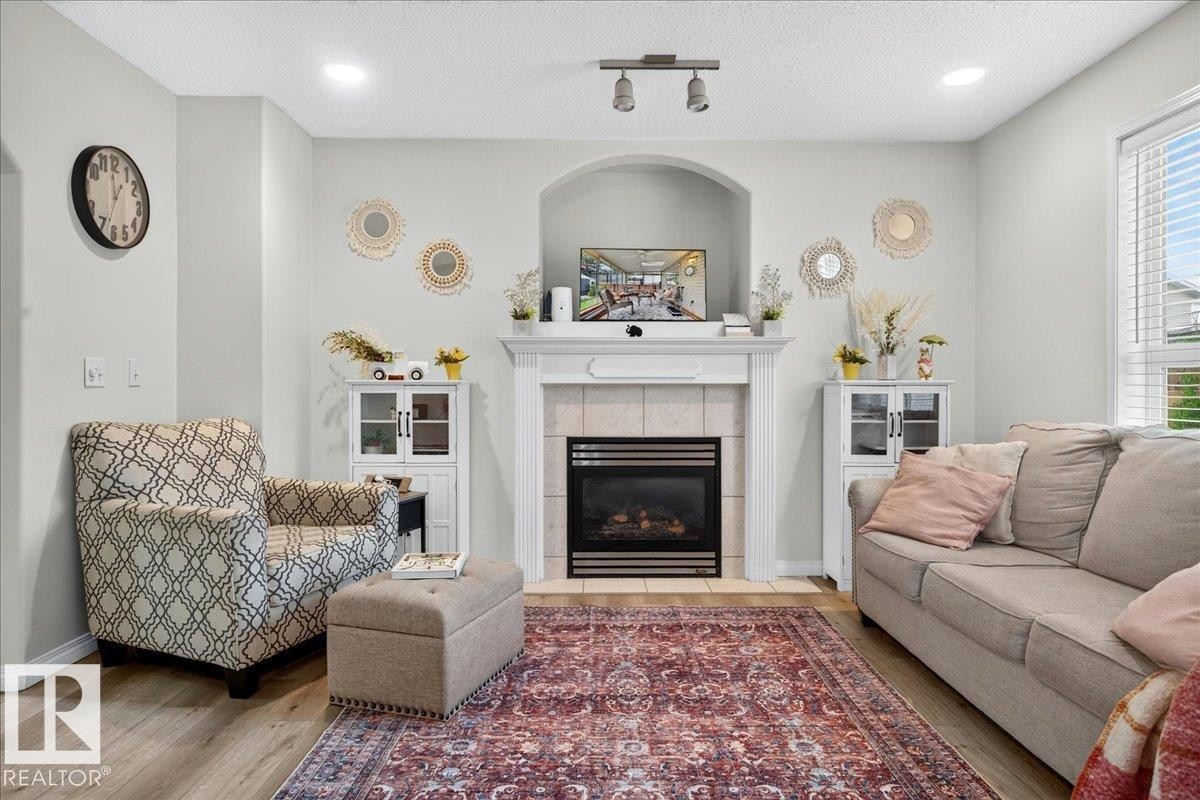 Living area featuring a fireplace with a white mantel and tile surround, wood-finish flooring, recessed lighting, a large window with blinds, and light gray wall paint - 6 Norfolk Close, St. Albert, AB - Indoor Photo Showing Living Room With Fireplace