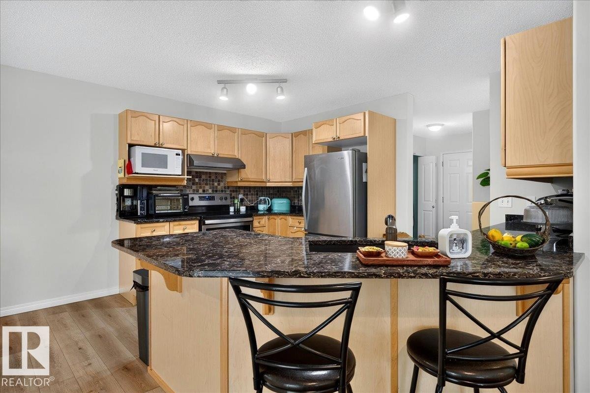 Kitchen featuring a breakfast bar with stone-finish countertop, light wood cabinetry, stainless steel appliances, and wood-finish flooring - 6 Norfolk Close, St. Albert, AB - Indoor Photo Showing Kitchen