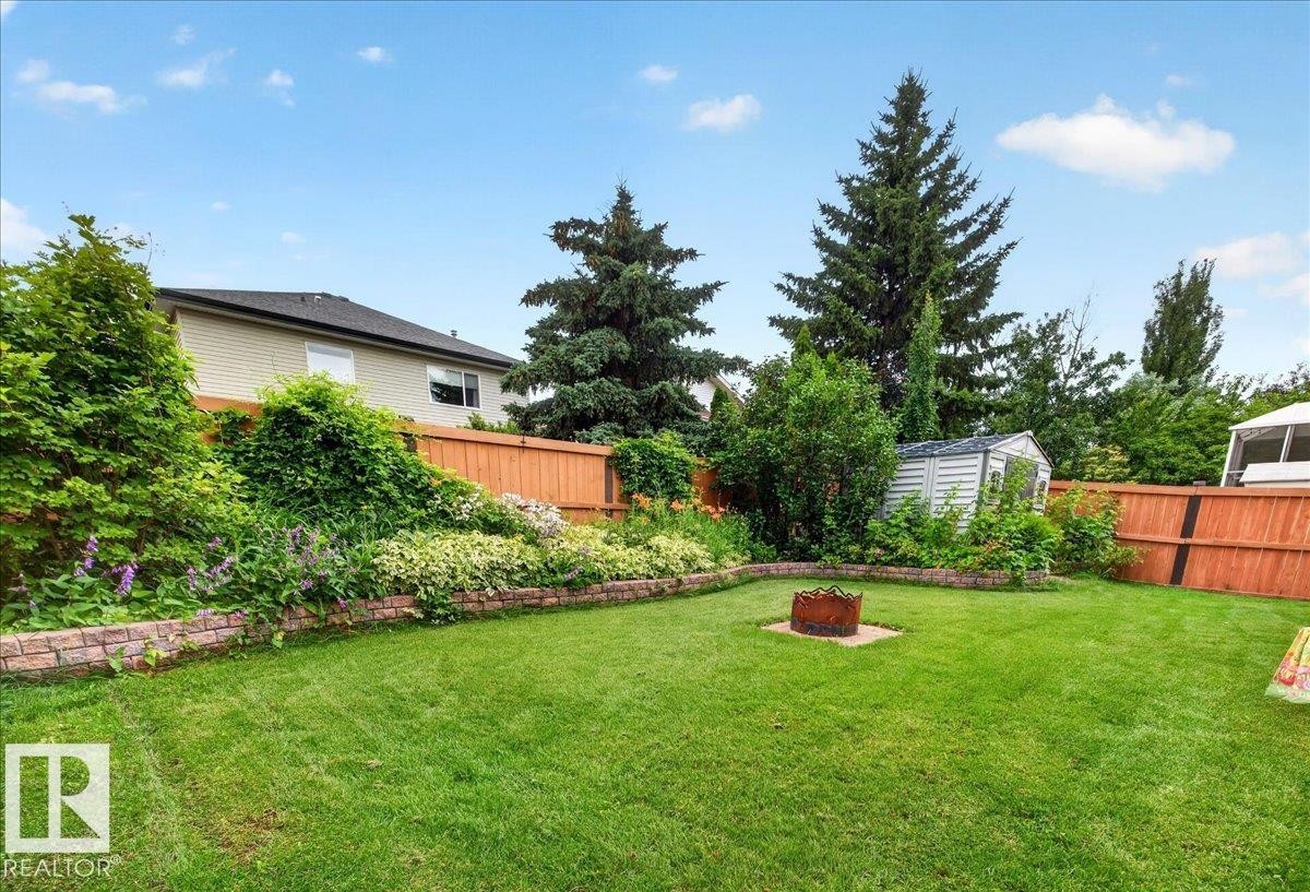 Expansive lawn featuring a fire pit, mature perimeter trees, and a garden bed with retaining wall - 6 Norfolk Close, St. Albert, AB - Outdoor With Backyard