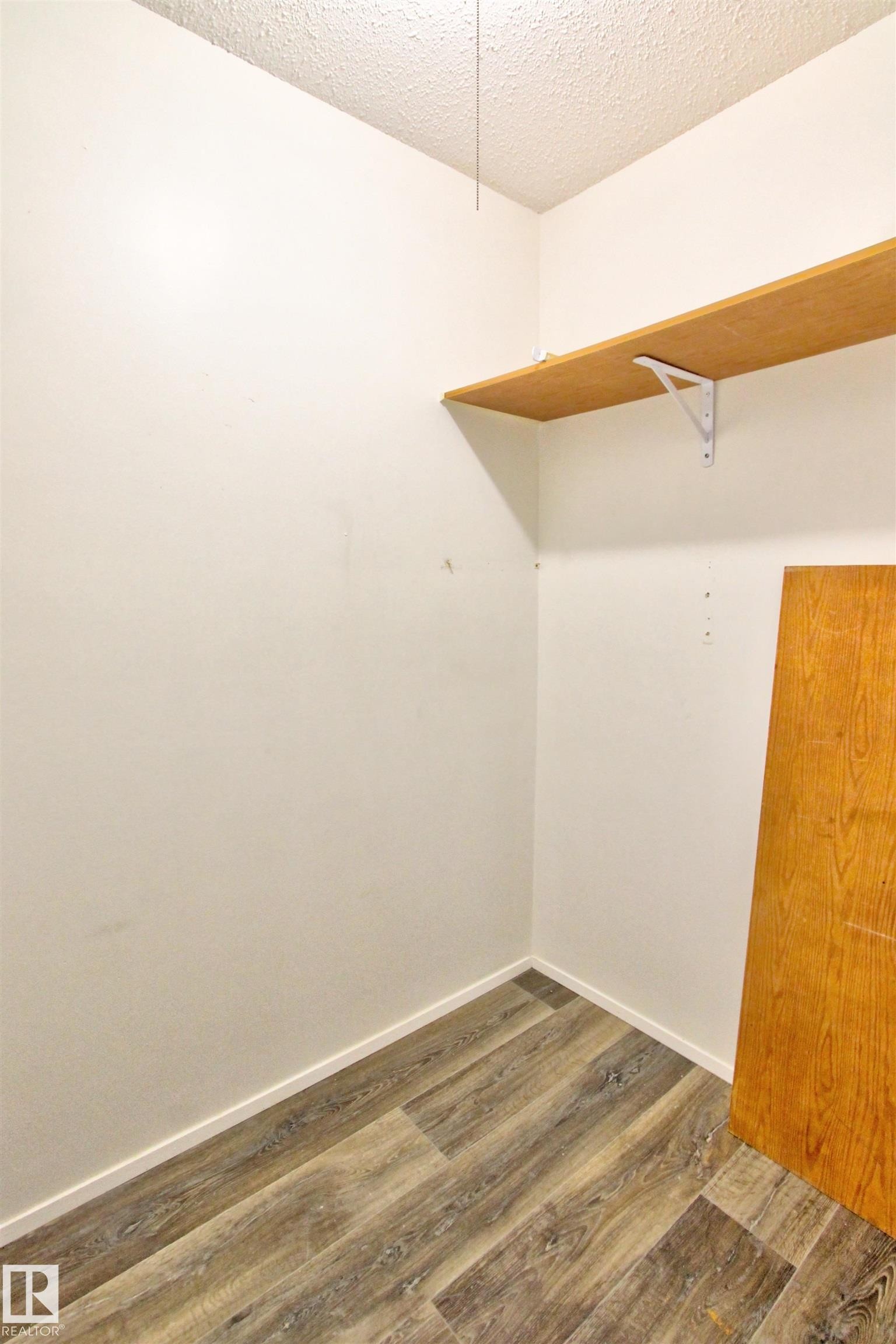 Corner closet featuring wood-finish flooring, a single wooden shelf with white brackets, white baseboards, and textured ceiling - #302 47 Akins Dr, St. Albert, AB - Indoor