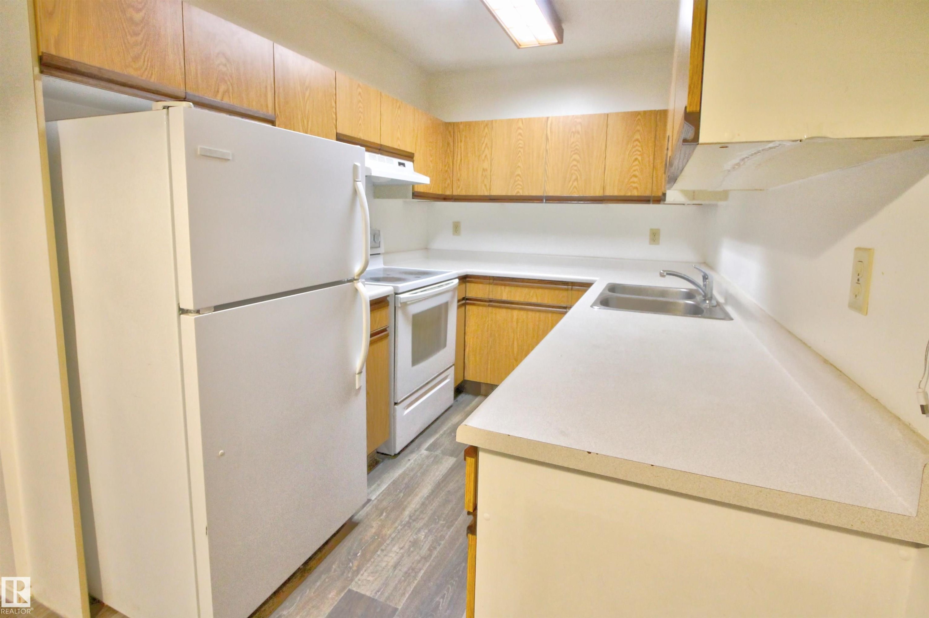 Compact kitchen featuring wood-finish cabinetry, white countertops, a double basin stainless steel sink, and wood-finish flooring - #302 47 Akins Dr, St. Albert, AB - Indoor Photo Showing Kitchen With Double Sink