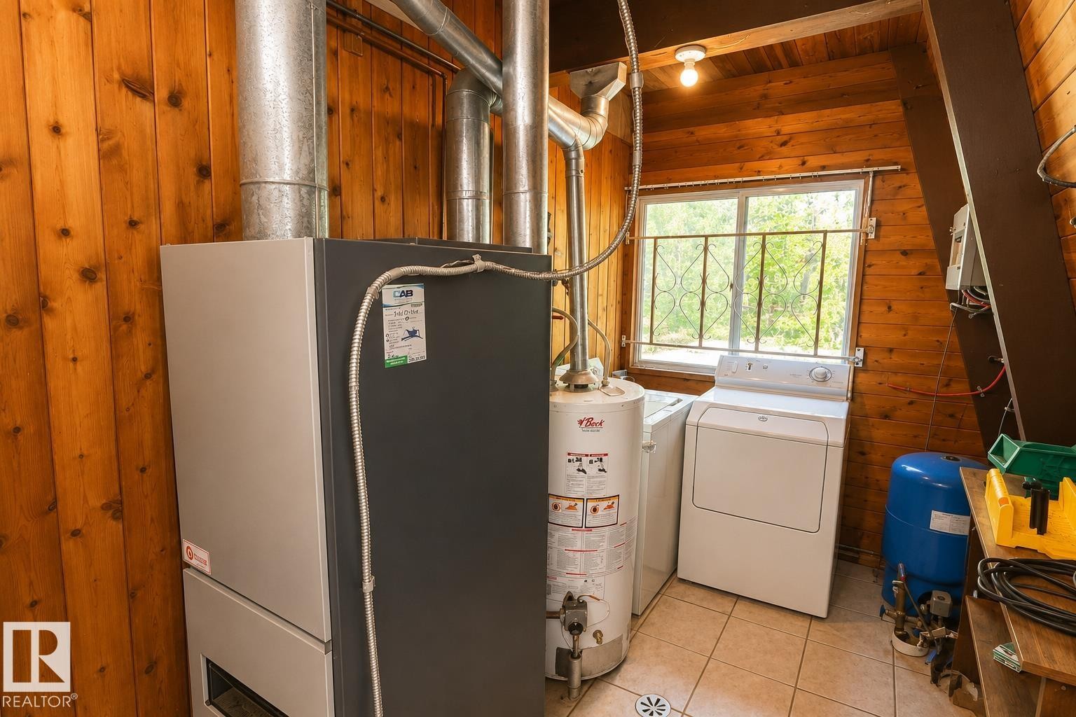 Dedicated utility area featuring wood-paneled walls, tile flooring, a window with decorative ironwork, and included washer, dryer, and water heater - 329 60417 Range Road 124, Rural St. Paul County, AB - Indoor Photo Showing Laundry Room