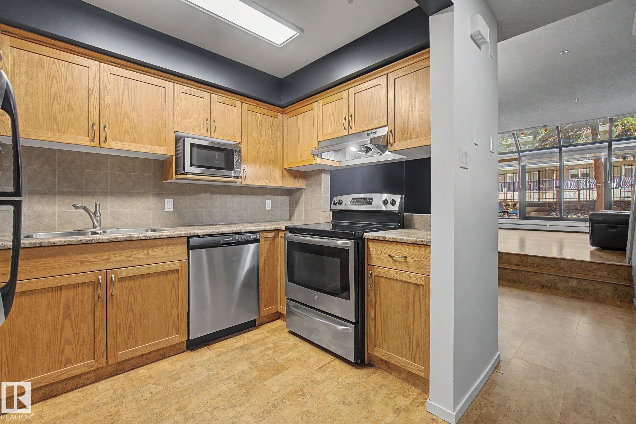 Kitchen featuring light wood cabinetry, stone-look countertops, and stainless steel appliances including a range, dishwasher, and microwave - 11150 83 Avenue Nw, Edmonton, AB
