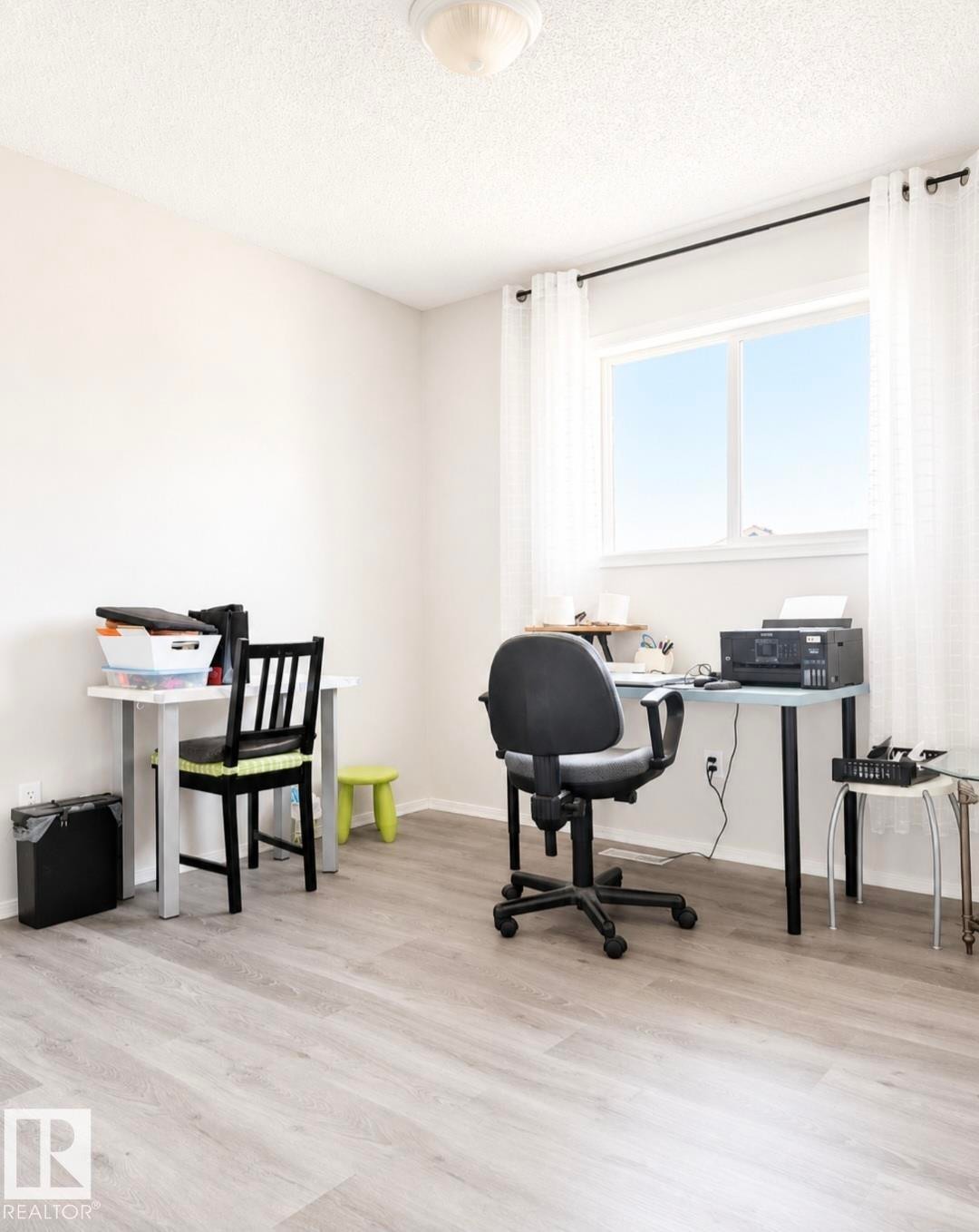 Bright interior room featuring a large window with white curtains, light wood-finish flooring, a neutral wall color, and a ceiling-mounted light fixture - 3236 27 Avenue Nw, Edmonton, AB - Indoor