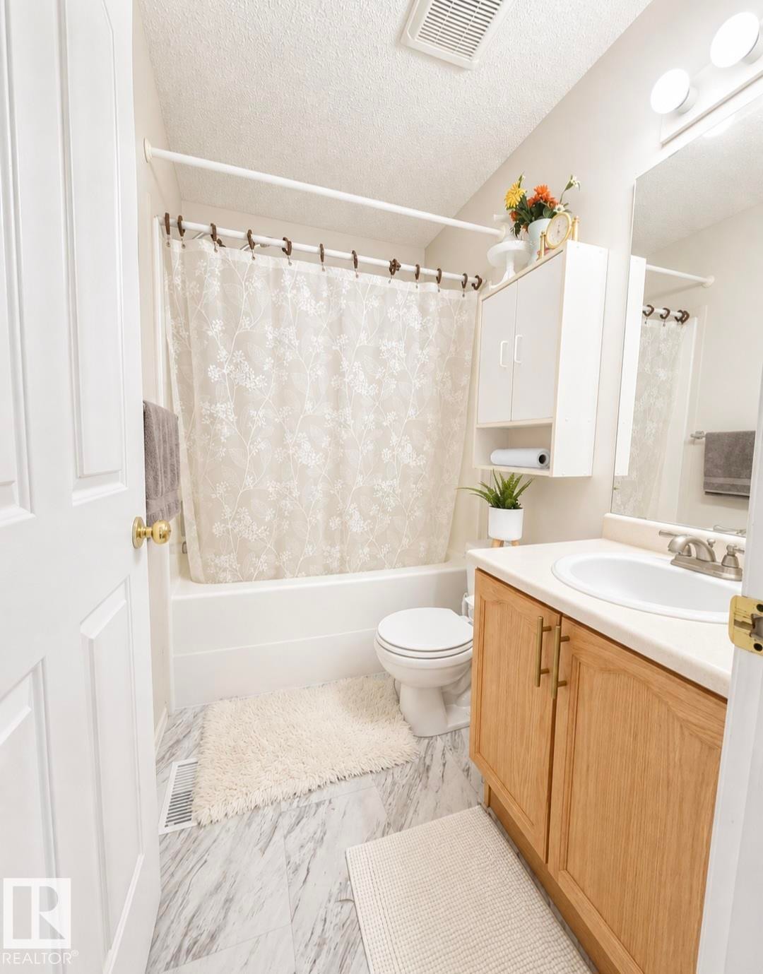 White bathtub with shower, toilet, wood-finish vanity with countertop and integrated sink, wall-mounted cabinet with open shelving, and patterned tile flooring - 3236 27 Avenue Nw, Edmonton, AB - Indoor Photo Showing Bathroom