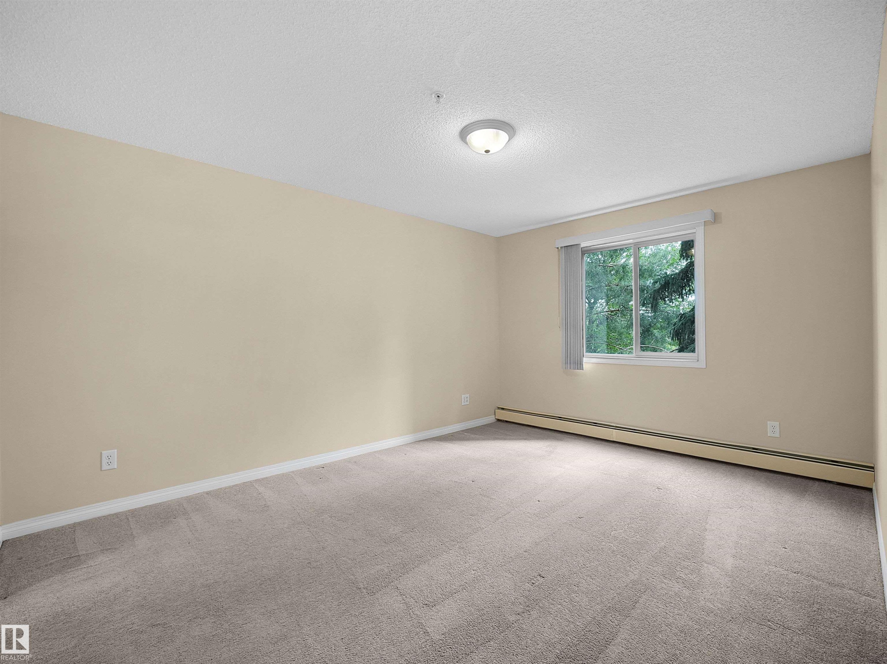 Spacious room featuring neutral carpeting, light-toned walls, and a large window with vertical blinds - #220 245 Edwards Dr Sw, Edmonton, AB - Indoor Photo Showing Other Room