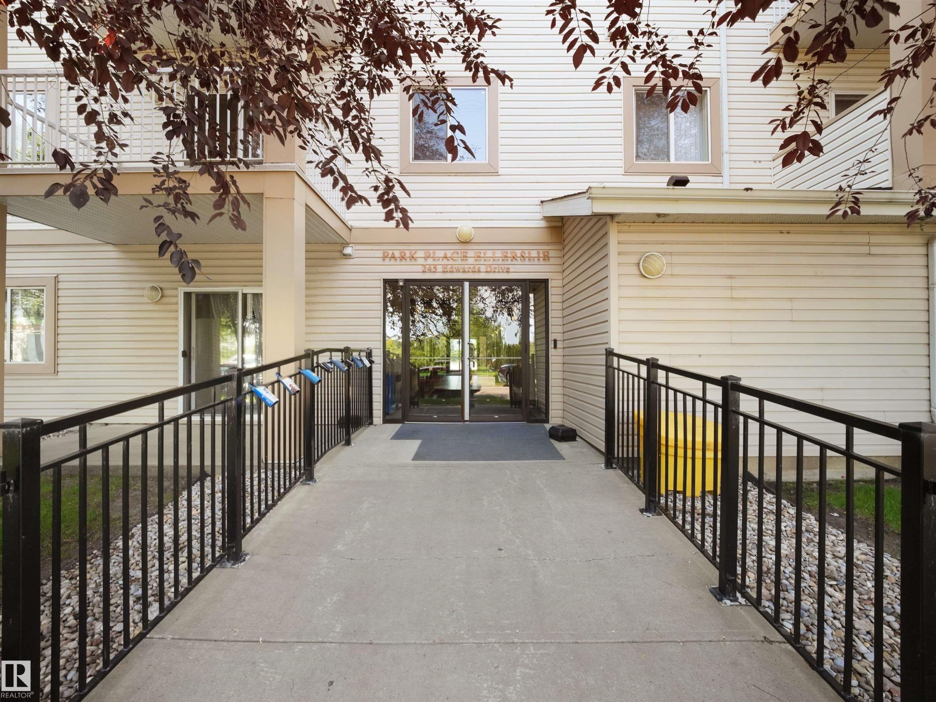 Building entrance featuring a concrete ramp with black metal railings, leading to glass double doors - #220 245 Edwards Dr Sw, Edmonton, AB - Outdoor With Exterior