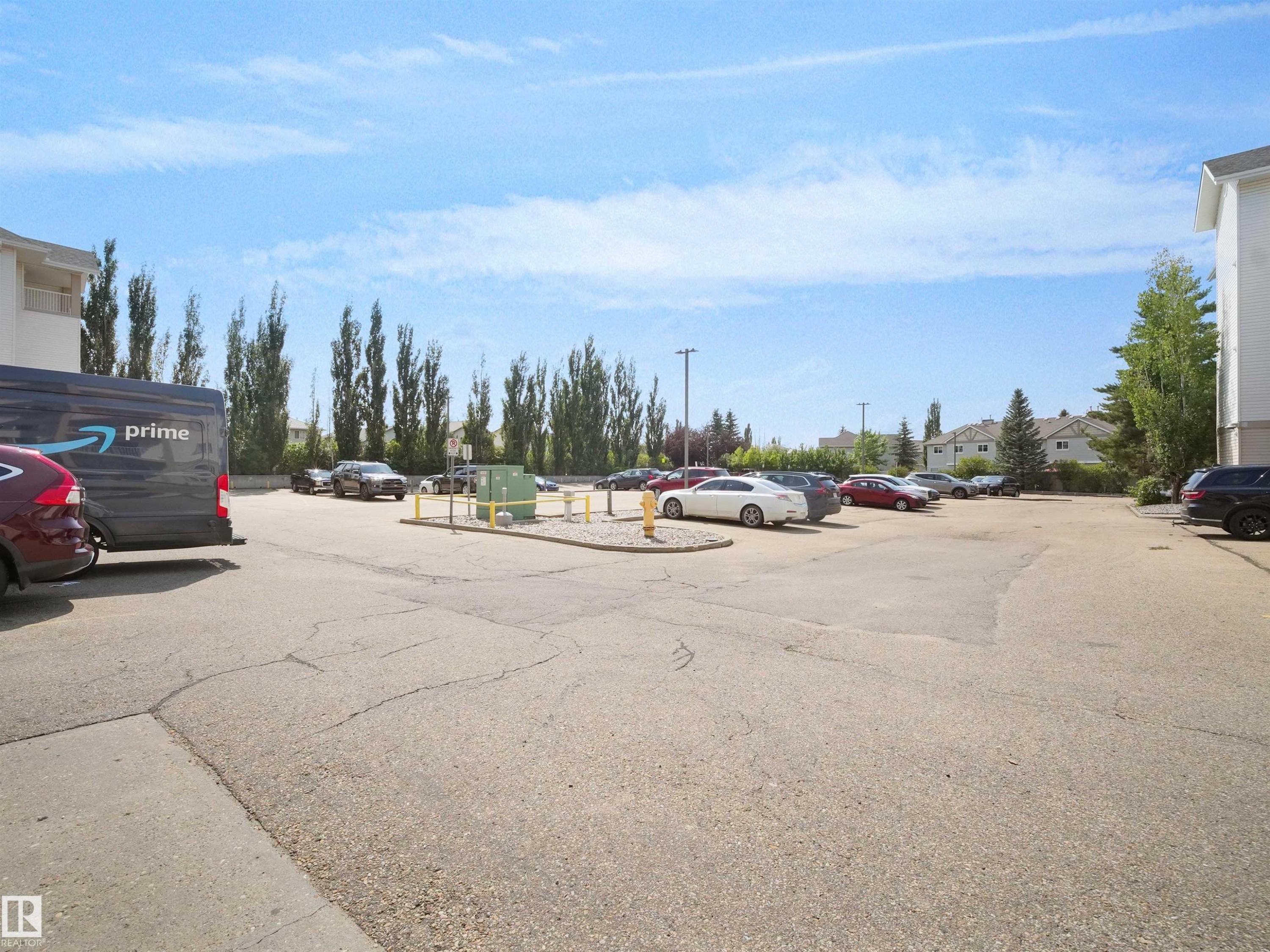 Paved parking area featuring multiple vehicle spaces, mature trees along the perimeter, and adjacent residential structures - #220 245 Edwards Dr Sw, Edmonton, AB - Outdoor
