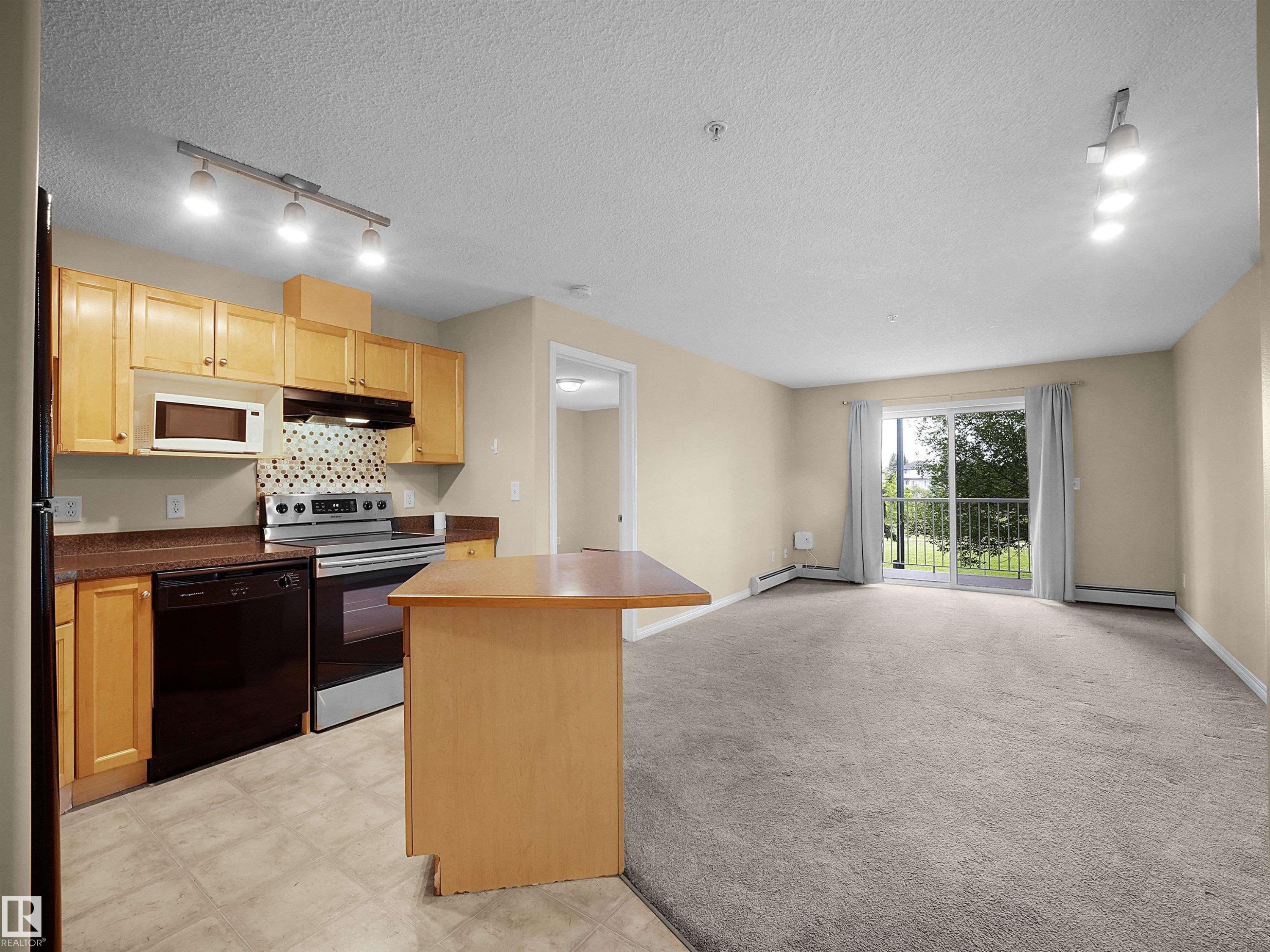 Open concept living area featuring a kitchen with wood cabinetry and a tiled backsplash, leading to a carpeted living space - #220 245 Edwards Dr Sw, Edmonton, AB - Indoor Photo Showing Kitchen With Double Sink