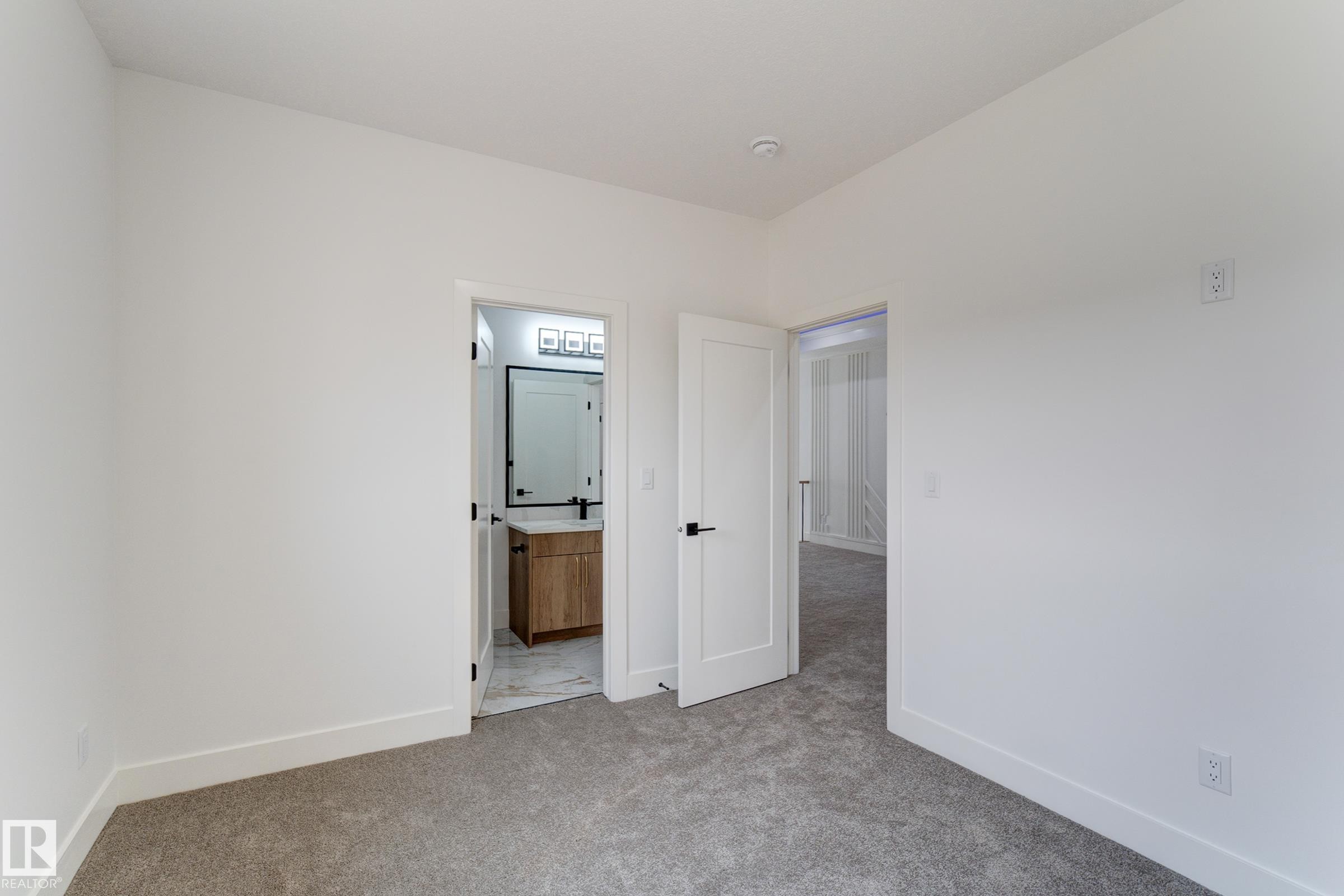 Neutral gray carpet flooring complements clean white walls in this room, featuring a doorway opening to a bathroom with a wood-finish vanity, a rectangular mirror with a matte black frame, and a three-light fixture - 4723 17 Avenue Sw, Edmonton, AB - Indoor Photo Showing Other Room