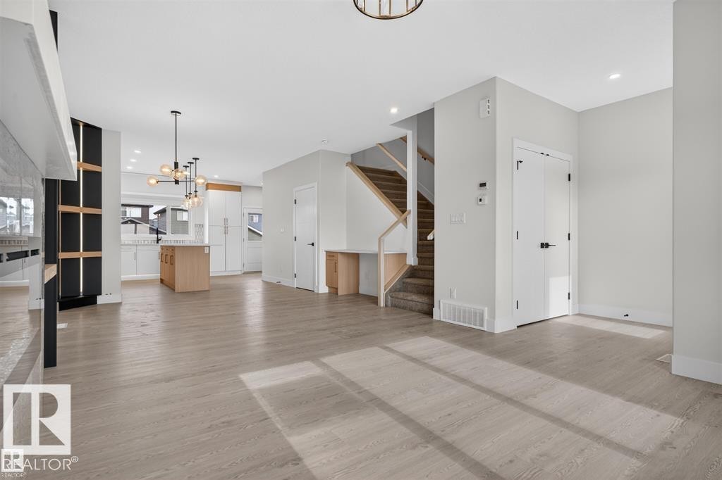 Spacious interior featuring light wood-finish flooring, a contemporary kitchen with an island, a modern chandelier, a built-in desk area, and a staircase with wood railing - 43 River Birch Way, St. Albert, AB - Indoor