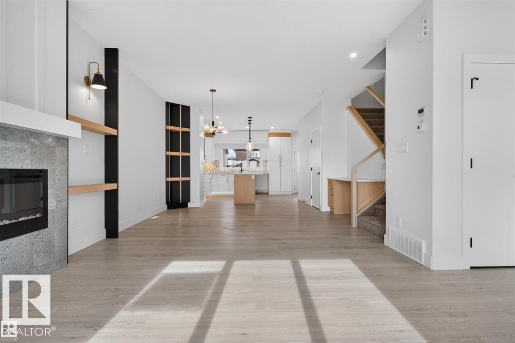 Open-concept living area featuring light wood-finish flooring, a modern fireplace with a grey stone surround, built-in shelving, a kitchen with an island, and a staircase with natural wood accents - 43 River Birch Way, St. Albert, AB - Indoor With Fireplace