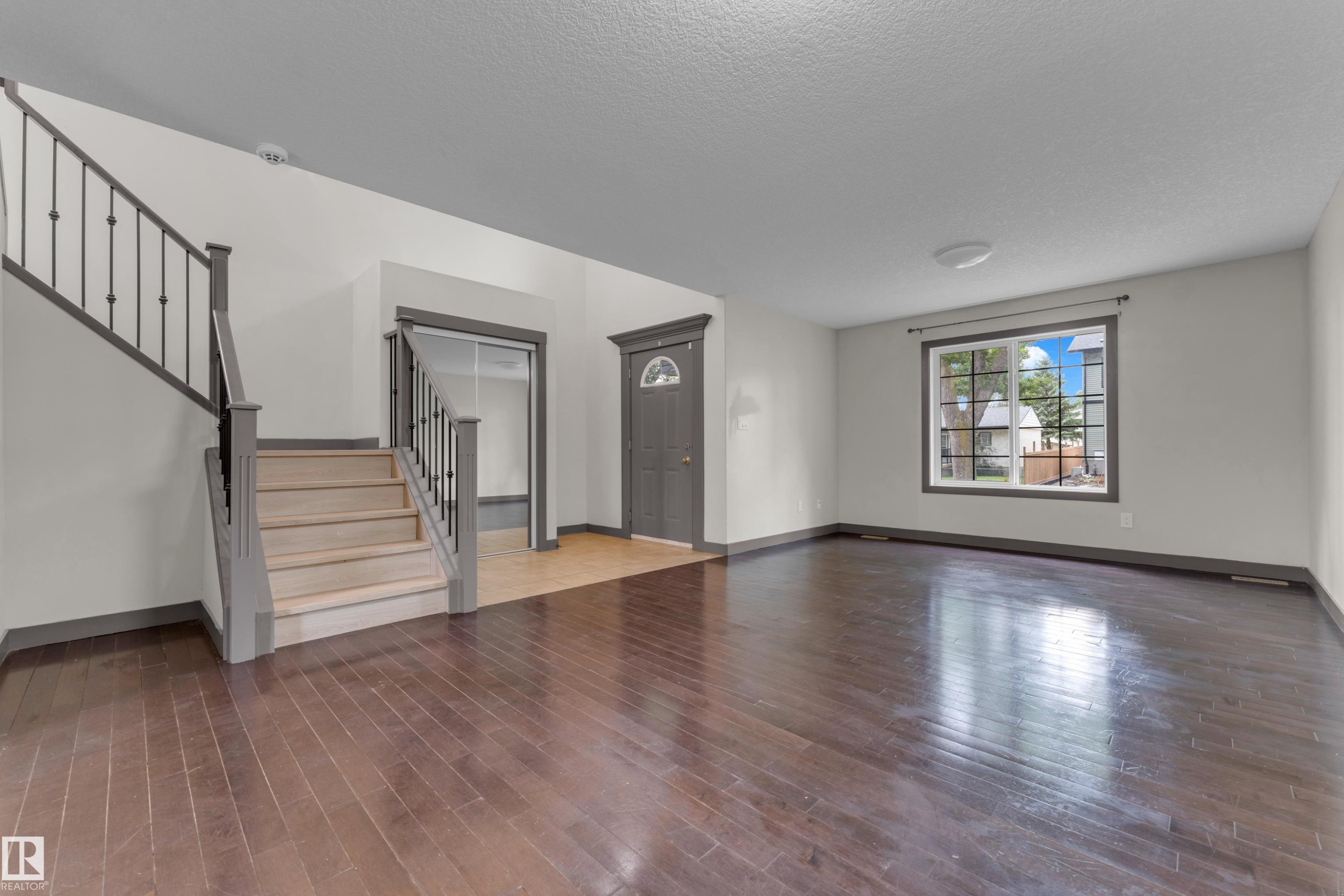 Spacious interior featuring dark wood-finish flooring, a prominent staircase with iron balusters, a front entry door with an arched window insert, a large multi-pane window, and light-toned walls with contrasting trim - 11817 37 Street Nw, Edmonton, AB - Indoor