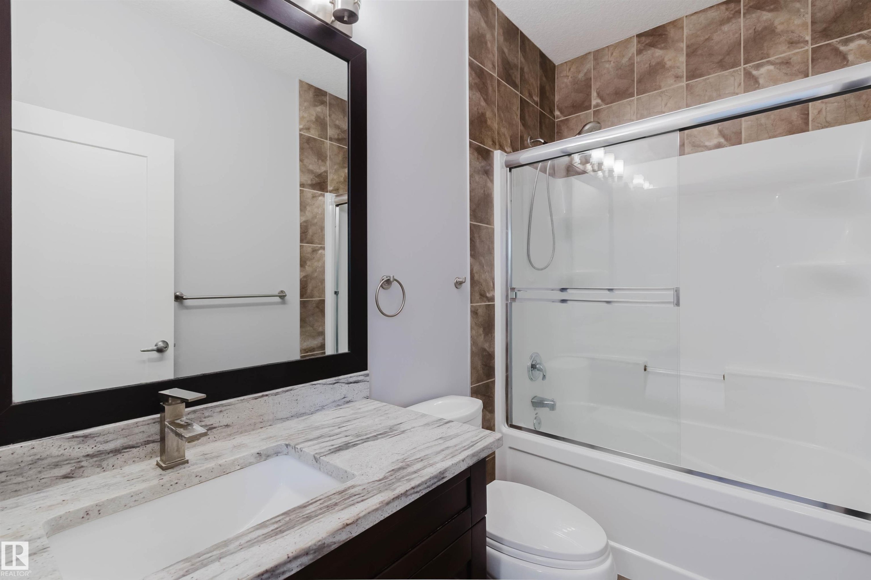 Bathroom featuring a combination tub and shower with a sliding glass enclosure, brown tiled shower surround, dark wood-finish vanity with an integrated sink, brushed nickel faucet, and a framed mirror - 10626 81 Avenue Nw, Edmonton, AB - Indoor Photo Showing Bathroom