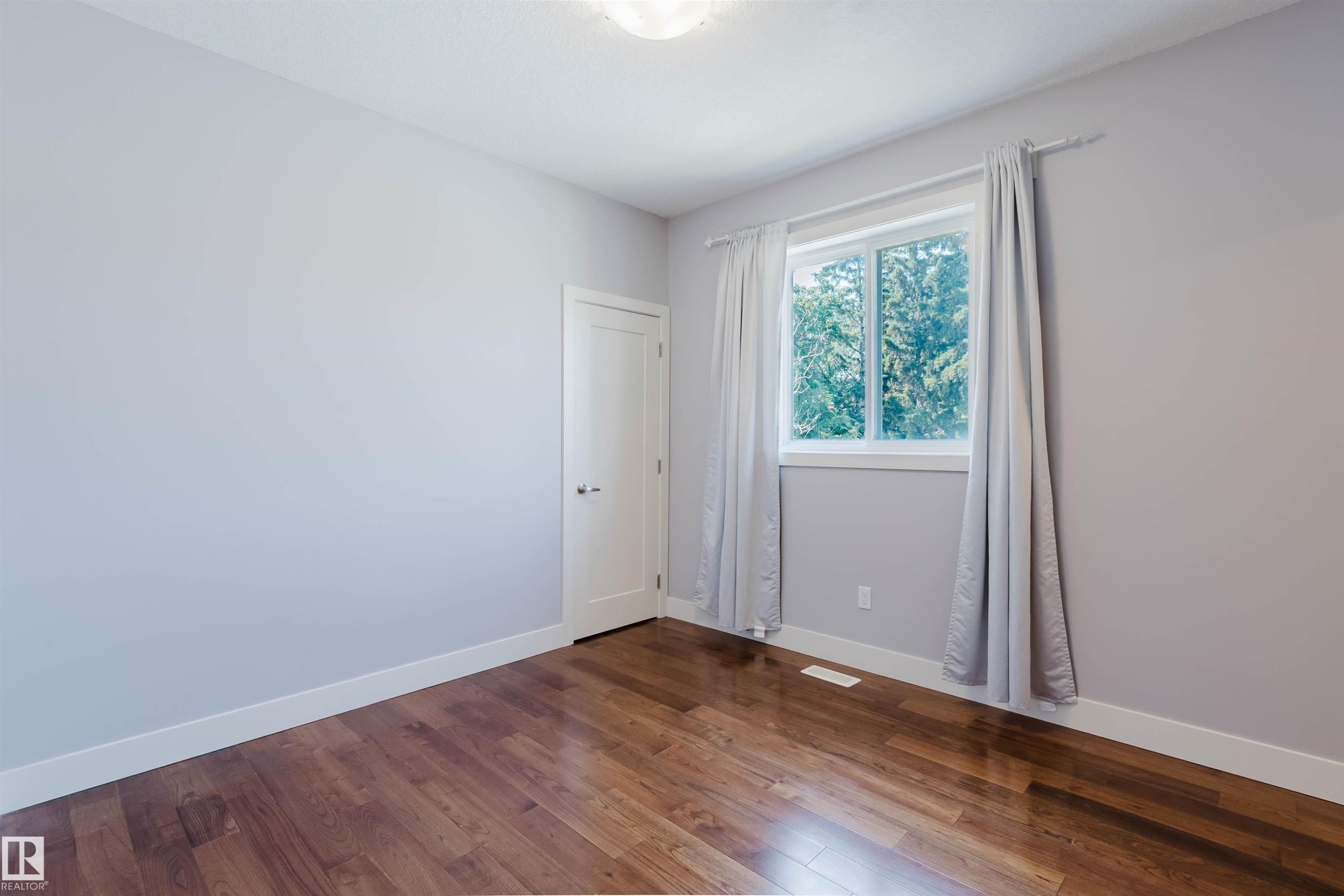 Spacious room featuring rich wood-finish flooring, light gray walls, white baseboards, and a white window frame - 10626 81 Avenue Nw, Edmonton, AB - Indoor Photo Showing Other Room