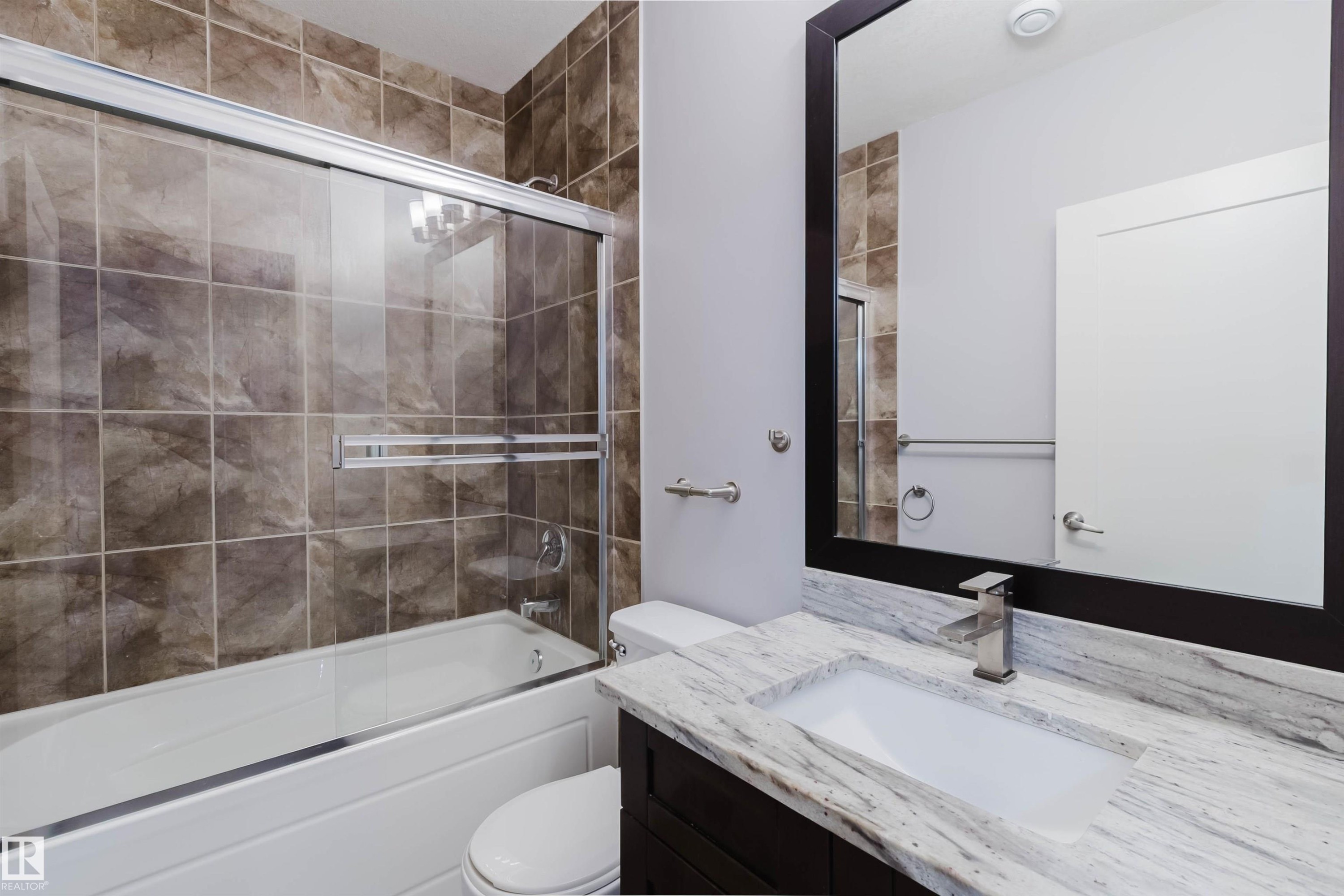Bathroom featuring a white bathtub with sliding glass doors, brown tiled shower surround, dark wood-finish vanity with an integrated rectangular sink, stone countertop, and a large framed mirror - 10626 81 Avenue Nw, Edmonton, AB - Indoor Photo Showing Bathroom