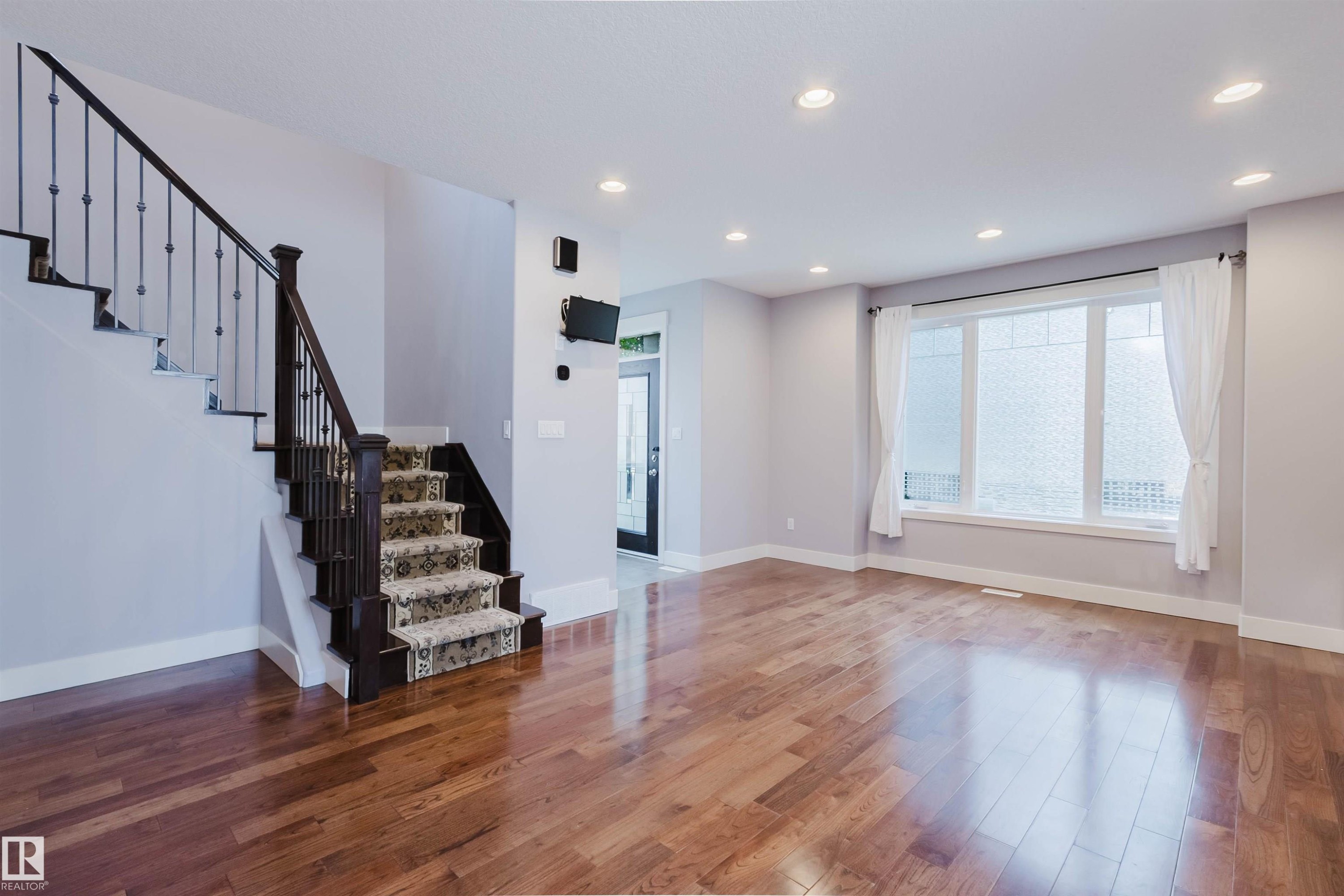 Spacious room featuring wood-finish flooring, a prominent staircase with dark wood newel post and wrought iron balusters, and a large multi-pane window - 10626 81 Avenue Nw, Edmonton, AB - Indoor Photo Showing Other Room