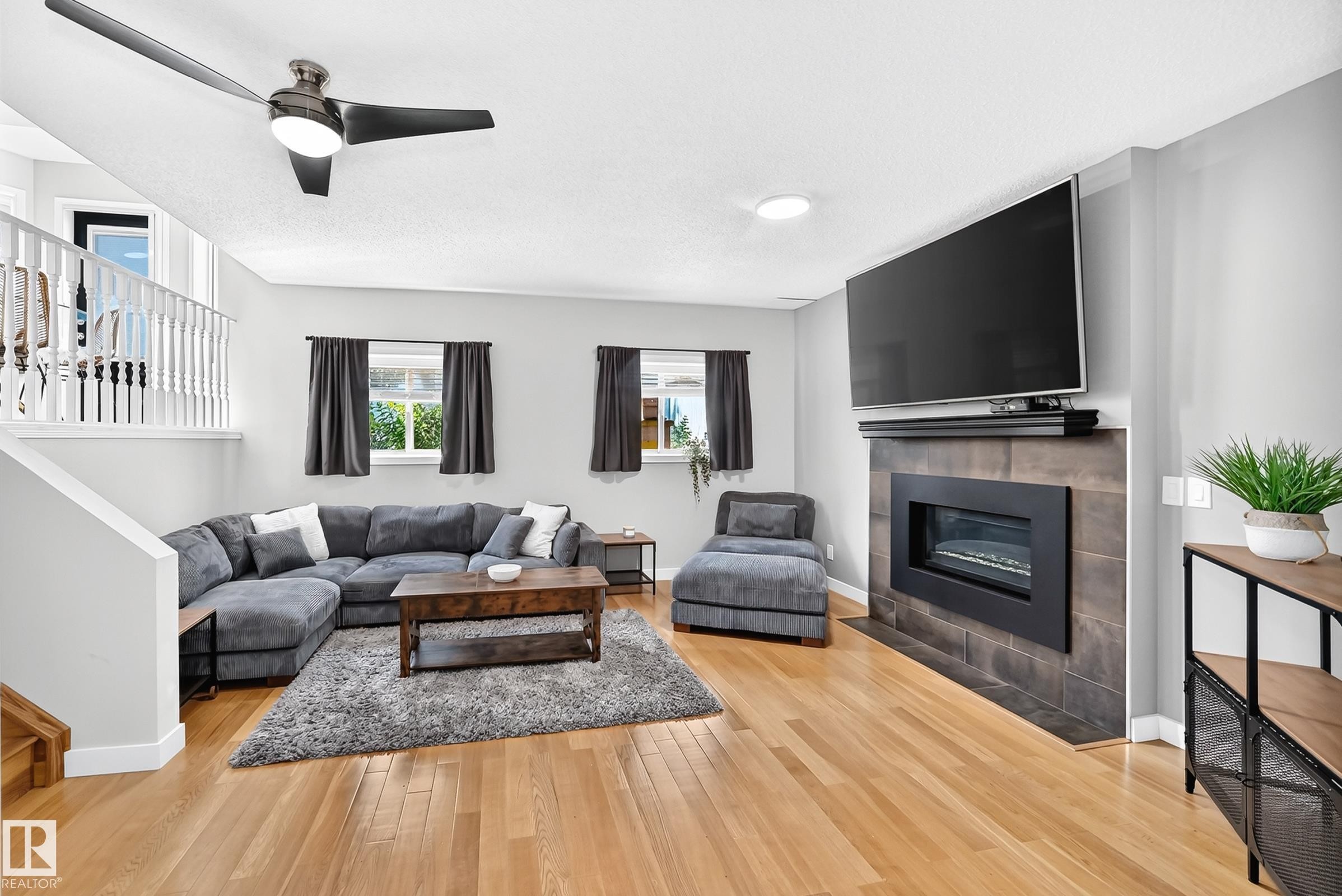 Bright living room featuring wood-finish flooring, a contemporary ceiling fan, and a fireplace with a dark tile surround - 3729 131A Avenue Nw, Edmonton, AB - Indoor Photo Showing Living Room With Fireplace