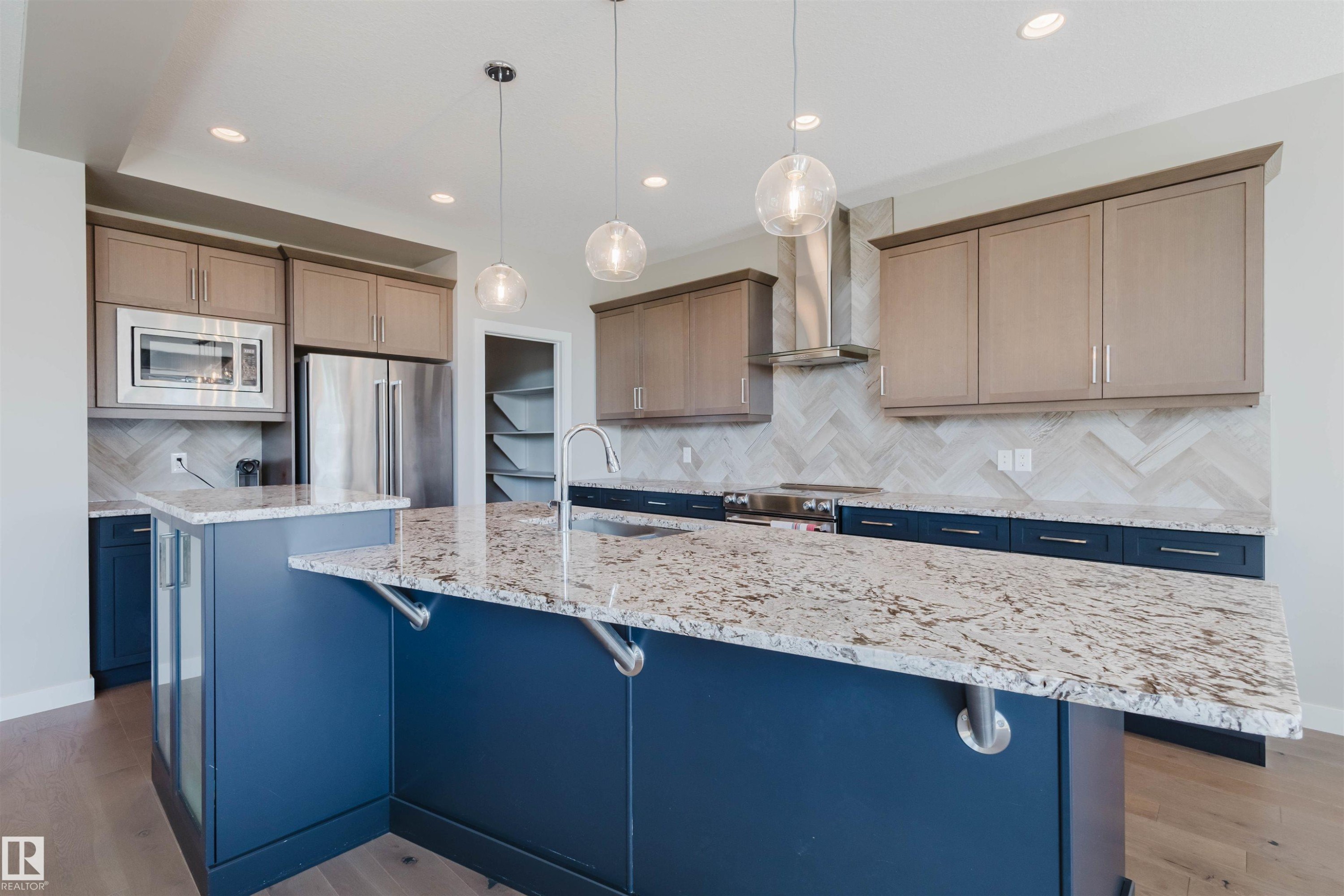 Kitchen featuring a large island with a speckled stone countertop, two-tone cabinetry, stainless steel appliances, a herringbone tile backsplash, and wood-finish flooring - 4125 Kennedy Green Sw, Edmonton, AB - Indoor Photo Showing Kitchen With Upgraded Kitchen