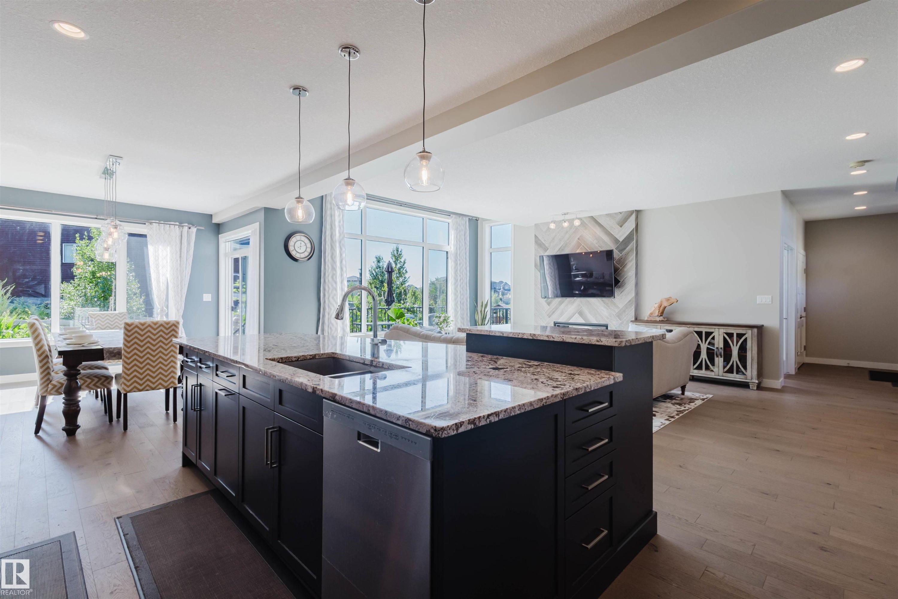 Expansive open-concept living area featuring a large kitchen island with a built-in sink and pendant lighting, wood-finish flooring, abundant natural light from large windows, and recessed ceiling lights - 4125 Kennedy Green Sw, Edmonton, AB - Indoor Photo Showing Kitchen With Upgraded Kitchen