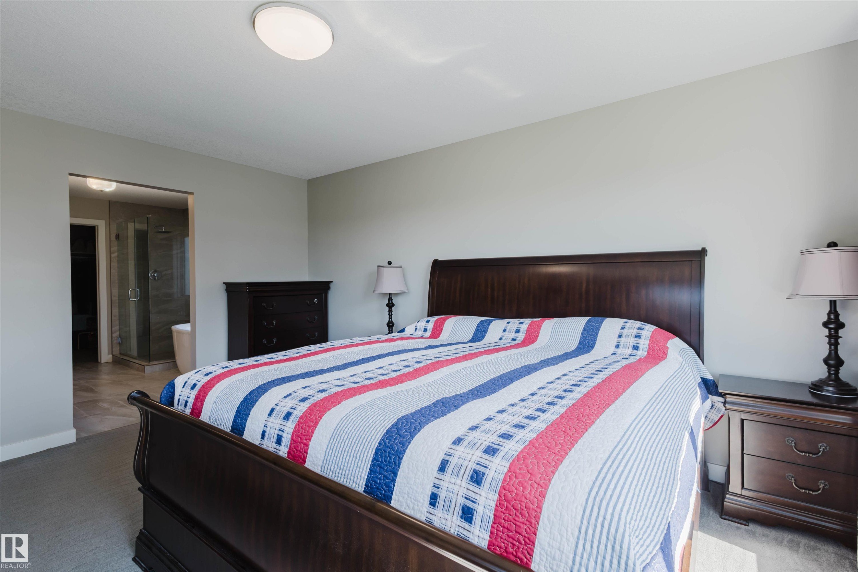 Carpeted bedroom with light gray walls, an oval flush mount ceiling light, and an open doorway revealing a bathroom with a glass-enclosed shower and a freestanding tub - 4125 Kennedy Green Sw, Edmonton, AB - Indoor Photo Showing Bedroom