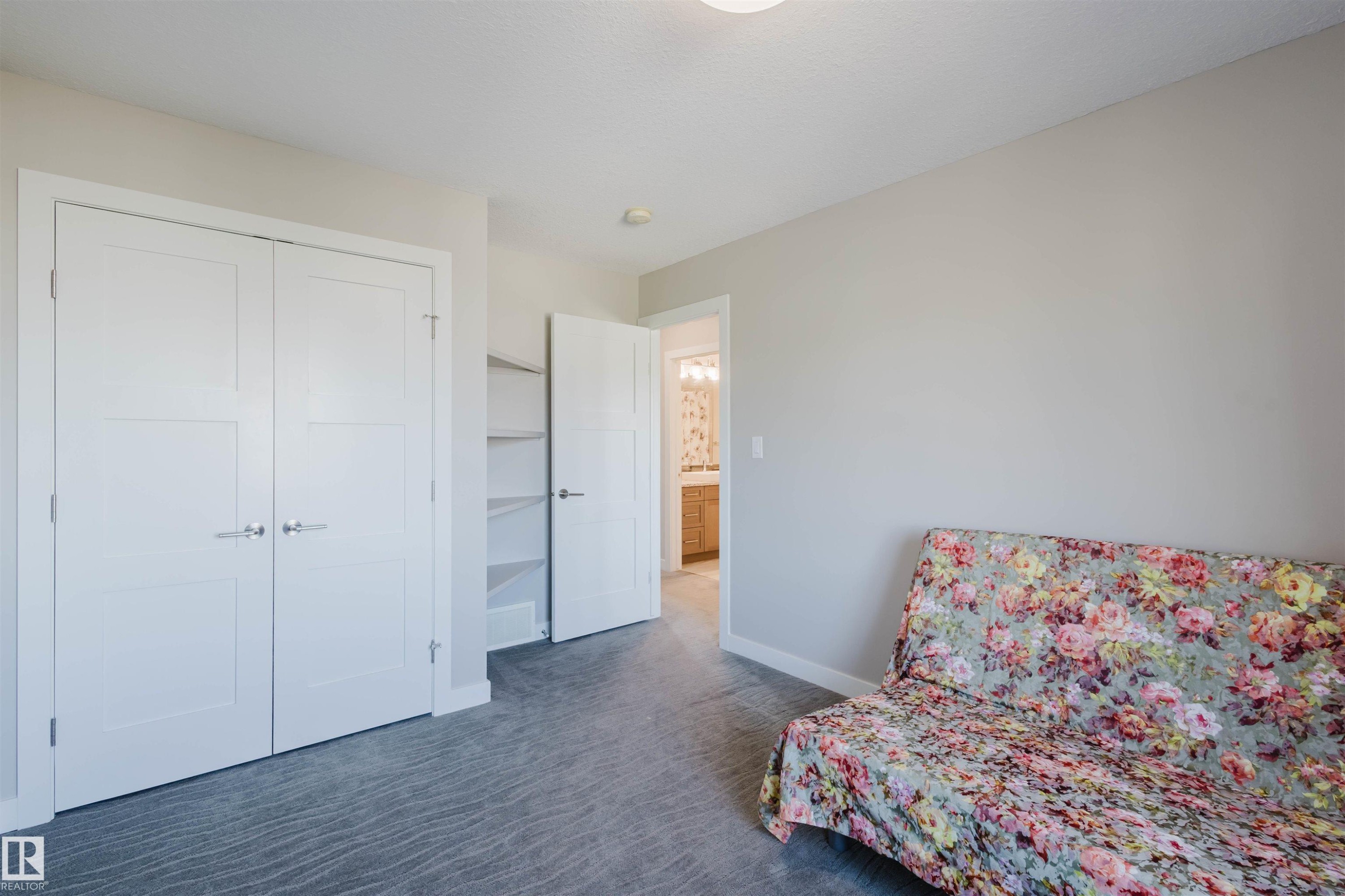 Carpeted room featuring a double-door closet with paneling and brushed nickel hardware, an open doorway revealing built-in shelving, and a second open doorway providing a glimpse into a bathroom with a wood-finish vanity - 4125 Kennedy Green Sw, Edmonton, AB - Indoor Photo Showing Bedroom