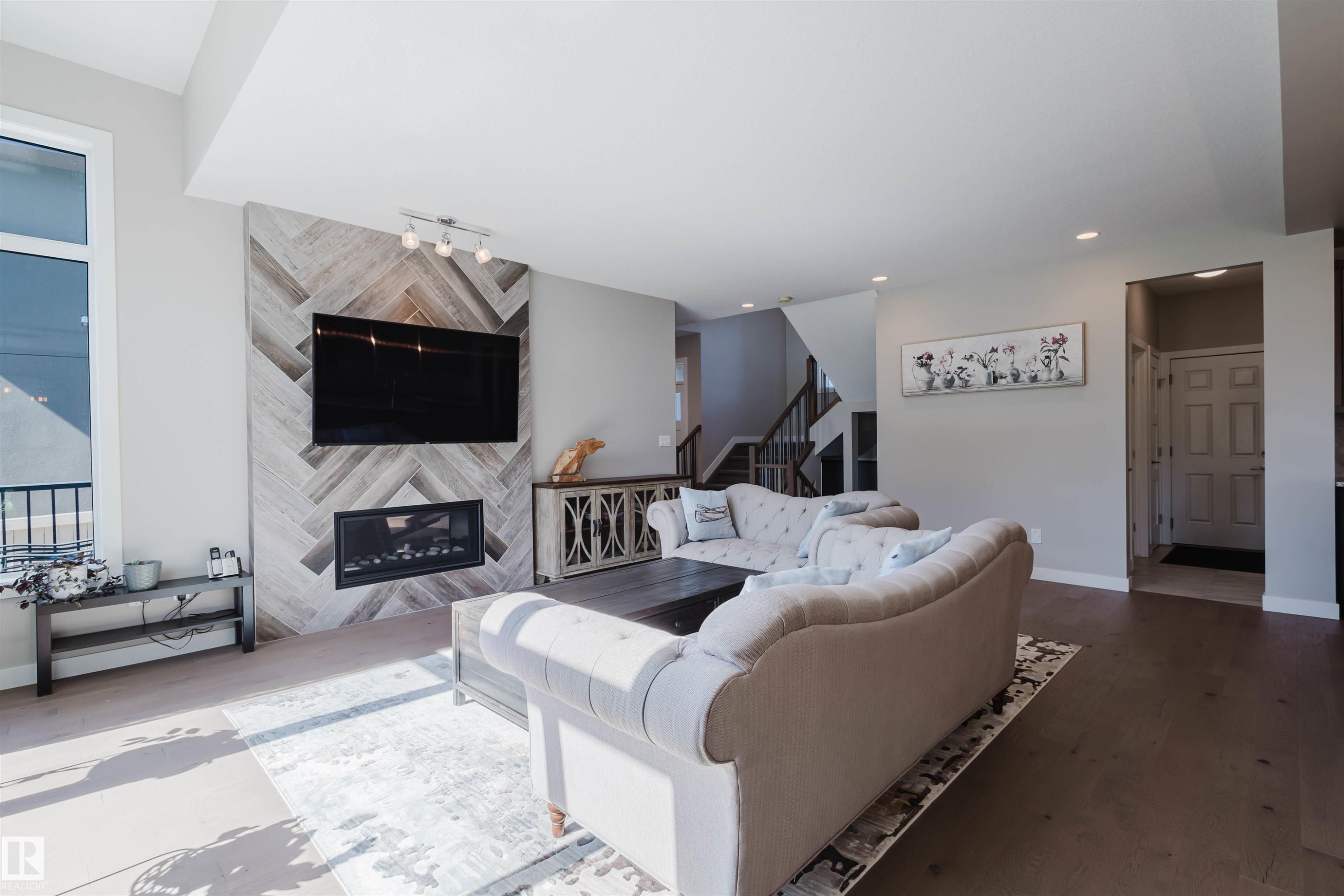 Living area featuring a fireplace with a herringbone tile surround, wall-mounted television, large window, and wood-finish flooring - 4125 Kennedy Green Sw, Edmonton, AB - Indoor Photo Showing Living Room With Fireplace