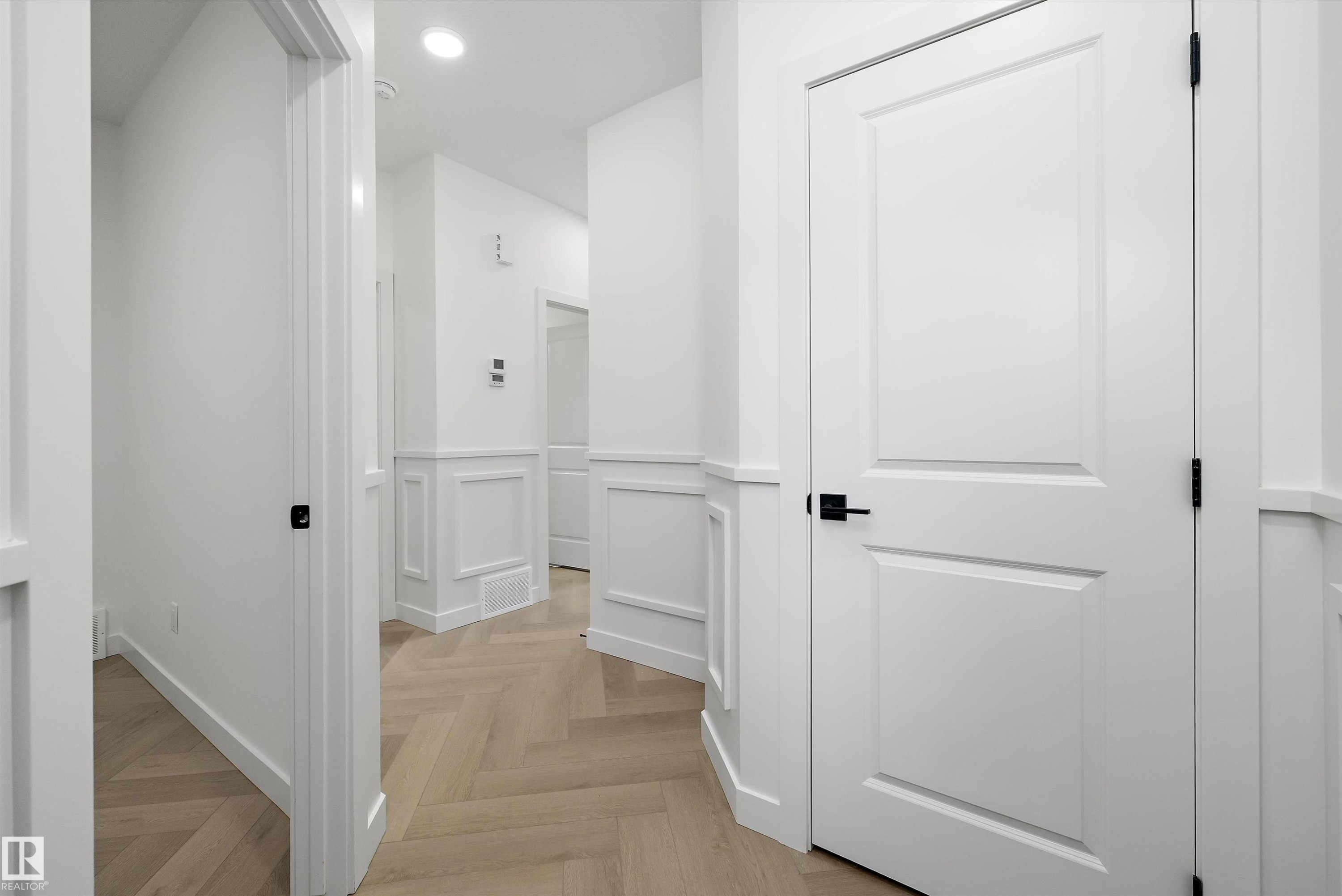 Elegant hallway featuring white shaker-style doors with matte black hardware, wainscoting paneling, recessed ceiling lighting, and warm wood-finish flooring in a herringbone pattern - 1776 Crossbill Loop Nw, Edmonton, AB
