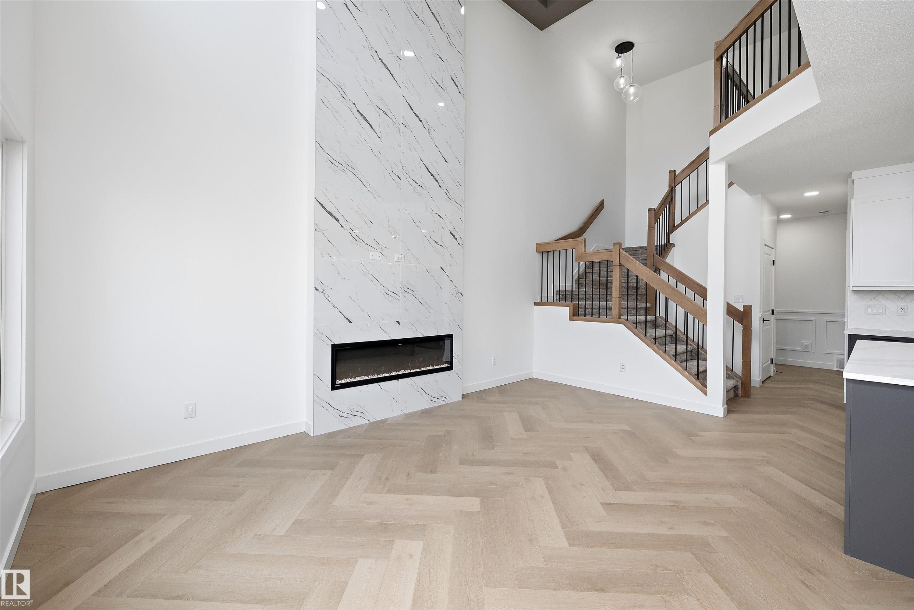 Herringbone wood-finish flooring, a tall fireplace with a marble-look surround, a multi-level staircase with wood railings and black spindles, and a modern pendant light fixture - 1776 Crossbill Loop Nw, Edmonton, AB