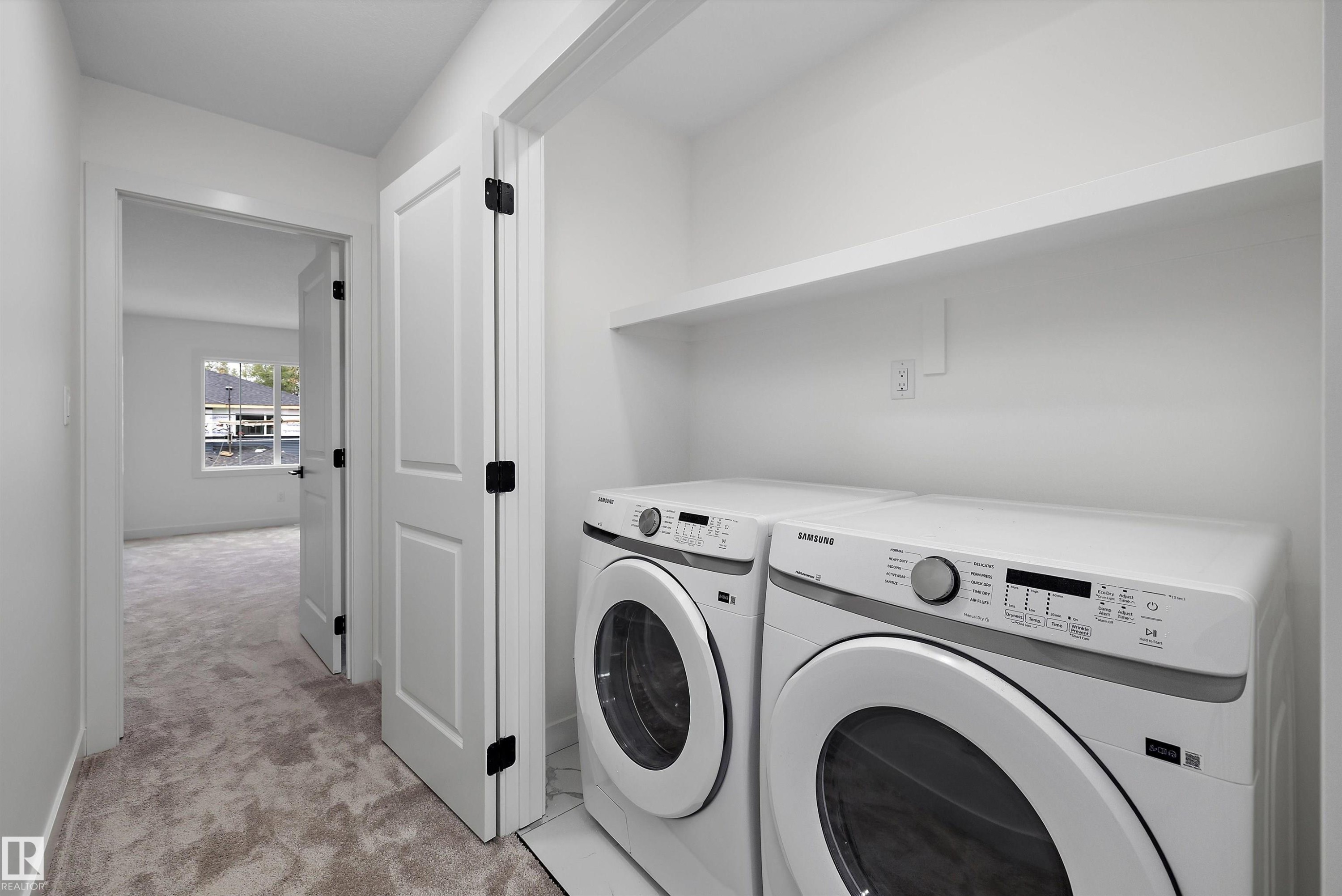 Modern laundry room featuring side-by-side front-load washer and dryer with stainless steel finishes, white shaker-style cabinetry, floating shelving above appliances, and neutral tile flooring throughout - 1776 Crossbill Loop Nw, Edmonton, AB