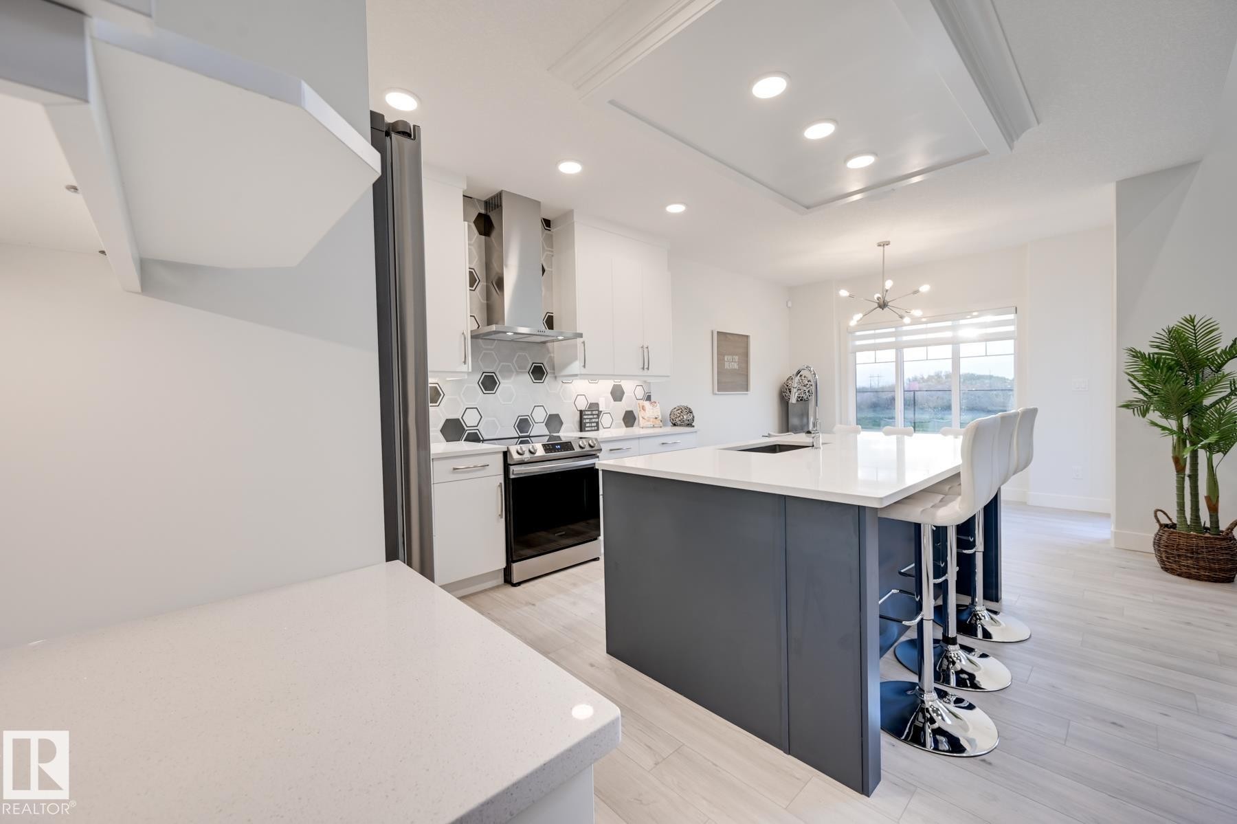 Modern kitchen featuring white cabinetry, hexagonal tile backsplash, stainless steel appliances, a center island with a white countertop, and wood-finish flooring - 1776 Crossbill Loop Nw, Edmonton, AB - Indoor Photo Showing Kitchen With Upgraded Kitchen
