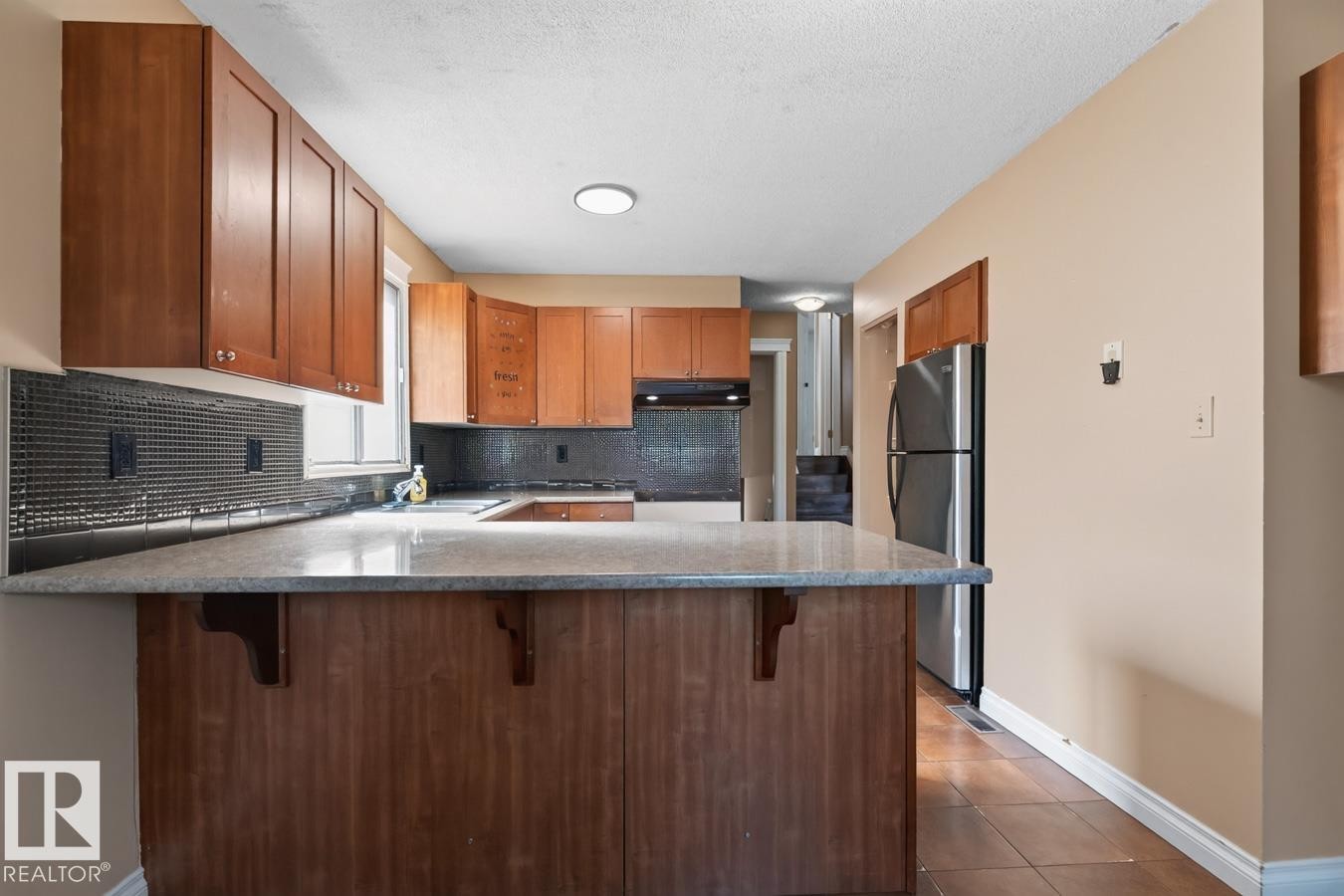 Kitchen featuring wood-finish cabinetry, a large peninsula with a light-toned countertop, and a dark tile backsplash - 4003 41 Avenue Nw, Edmonton, AB - Indoor Photo Showing Kitchen