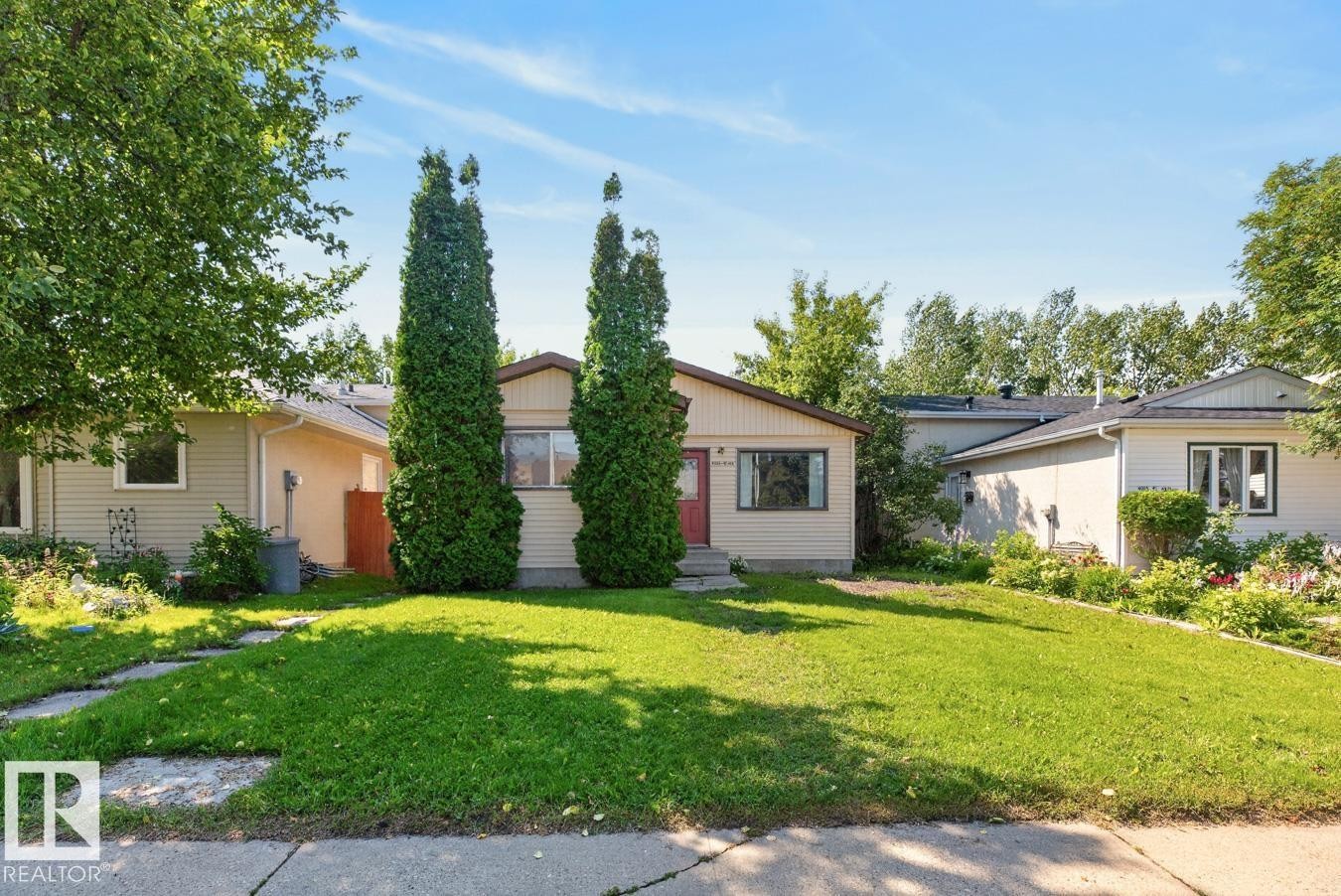 Ranch-style residence featuring beige siding, a red entry door, a prominent front lawn, two tall evergreen trees, and a concrete pathway leading to the entrance - 4003 41 Avenue Nw, Edmonton, AB - Outdoor