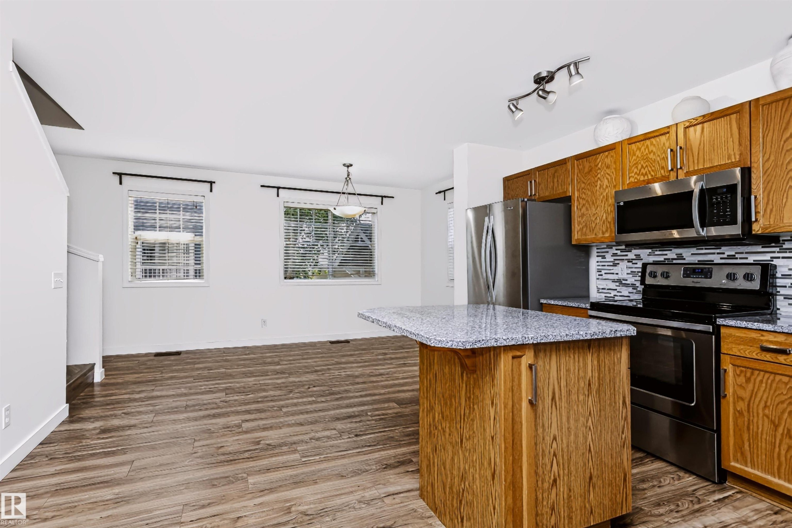 Kitchen featuring wood-finish cabinetry, stainless steel appliances, a mosaic tile backsplash, and a granite-look island with breakfast bar seating - #66 8304 11 Av Sw, Edmonton, AB - Indoor Photo Showing Kitchen With Stainless Steel Kitchen