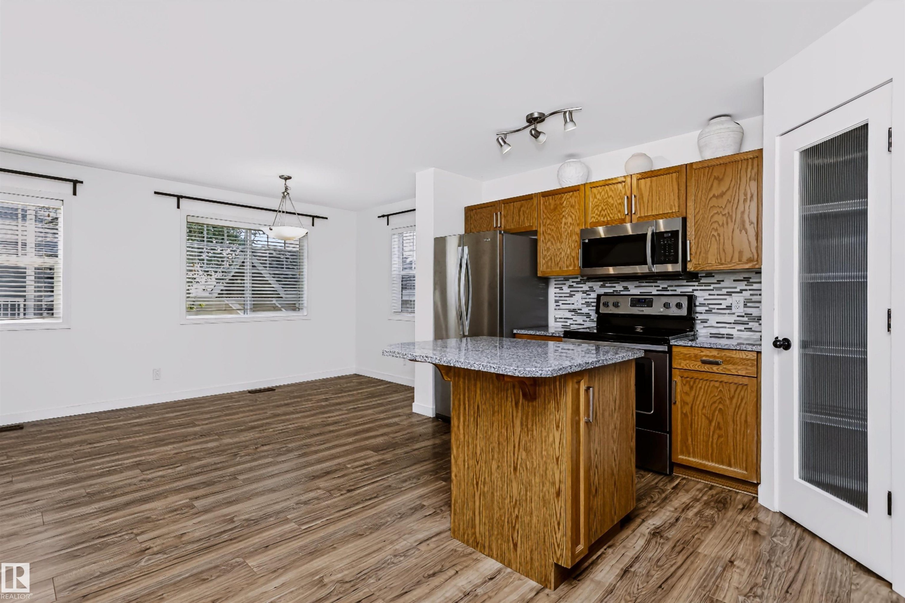 Open-concept kitchen and dining area featuring wood-finish flooring, a kitchen island with a speckled countertop, stainless steel appliances, and a tiled backsplash - #66 8304 11 Av Sw, Edmonton, AB - Indoor Photo Showing Kitchen