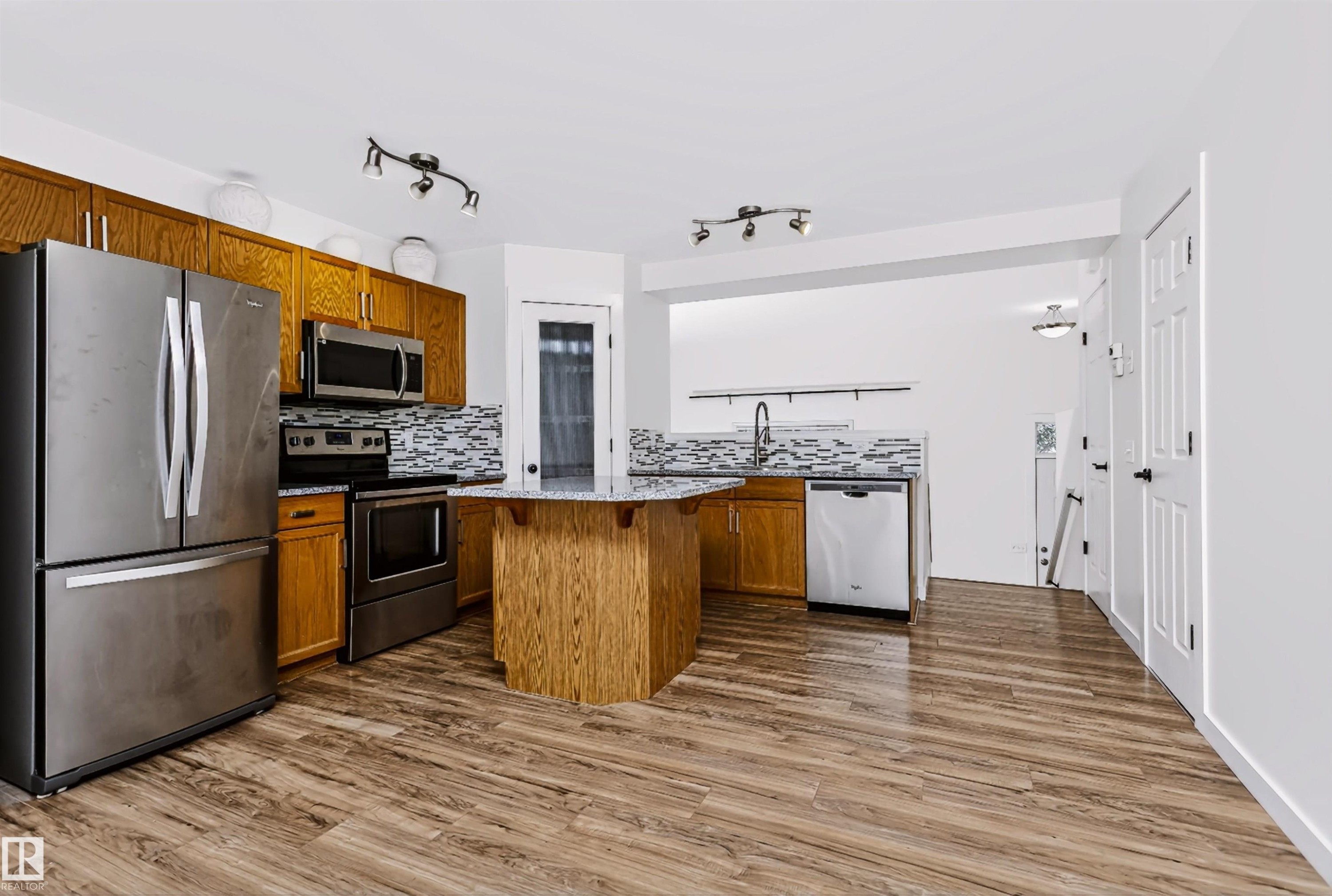 Kitchen featuring wood-finish flooring, a central island with a granite-style countertop, stainless steel appliances, a mosaic tile backsplash, and track lighting - #66 8304 11 Av Sw, Edmonton, AB - Indoor Photo Showing Kitchen With Stainless Steel Kitchen