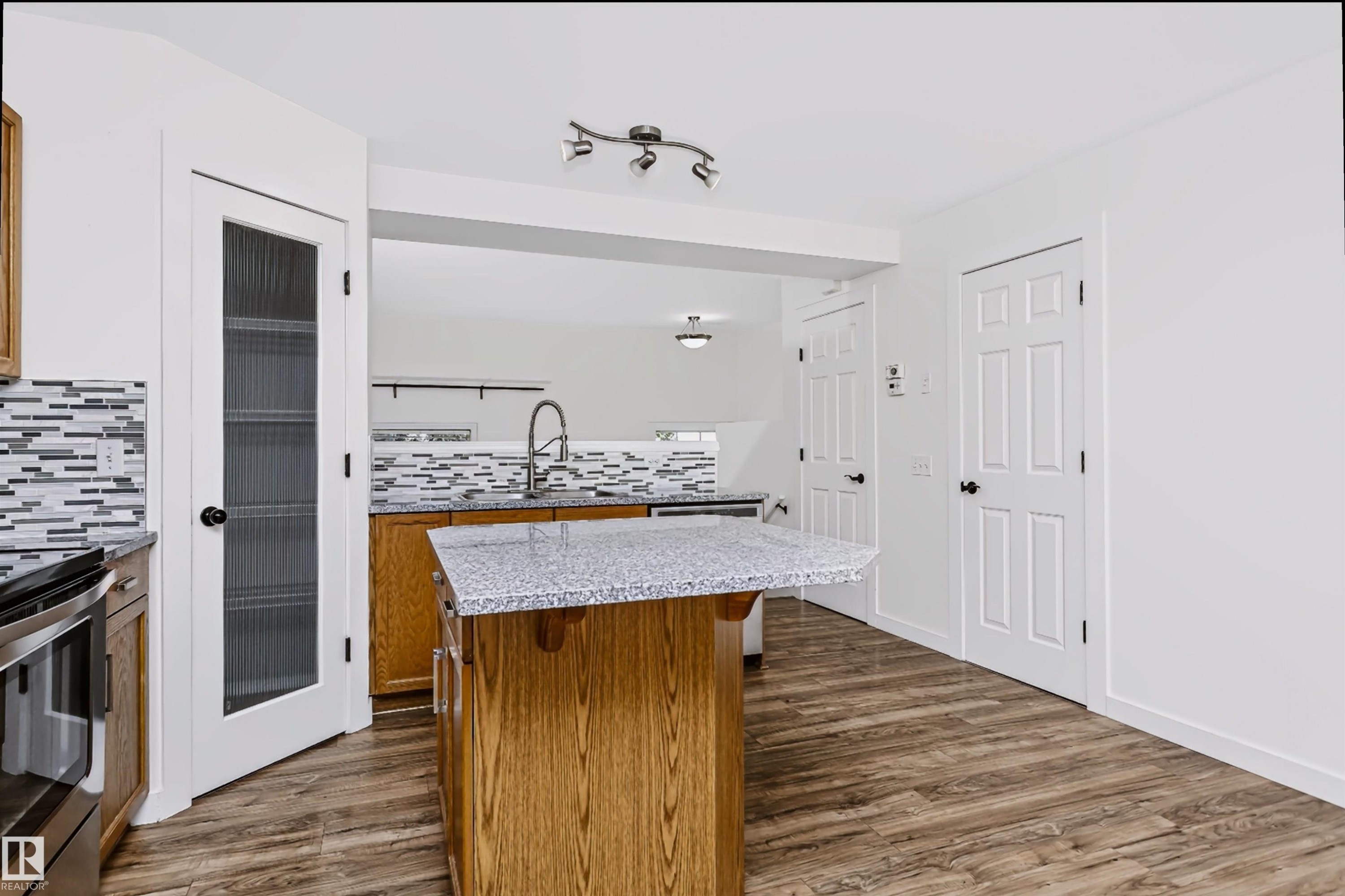 Kitchen featuring an island with a speckled countertop, wood-finish flooring, a mosaic tile backsplash, stainless steel appliances, and a glass-paneled pantry door - #66 8304 11 Av Sw, Edmonton, AB - Indoor Photo Showing Kitchen