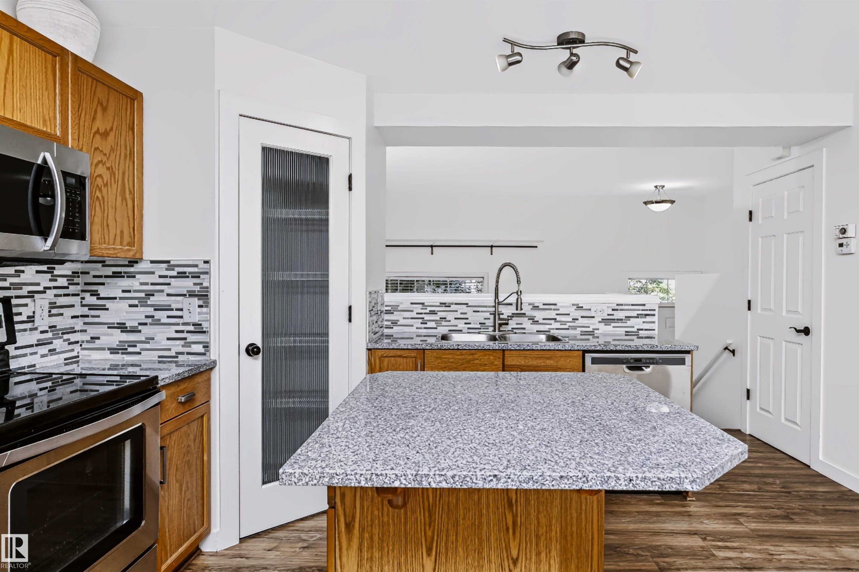 Kitchen featuring a multi-tone tile backsplash, stainless steel appliances, wood-finish flooring, and a central island with a granite-style countertop - #66 8304 11 Av Sw, Edmonton, AB - Indoor Photo Showing Kitchen With Double Sink With Upgraded Kitchen