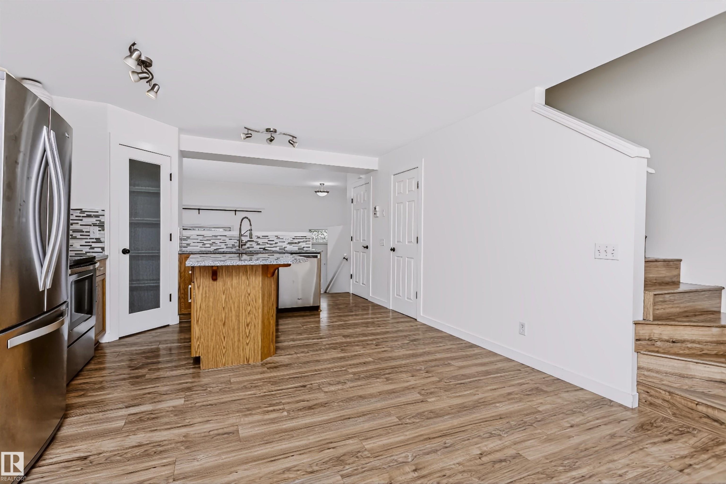 Open-concept kitchen and living area featuring wood-finish flooring, a kitchen island with a stone-look countertop, stainless steel appliances, and a tiled kitchen backsplash - #66 8304 11 Av Sw, Edmonton, AB - Indoor