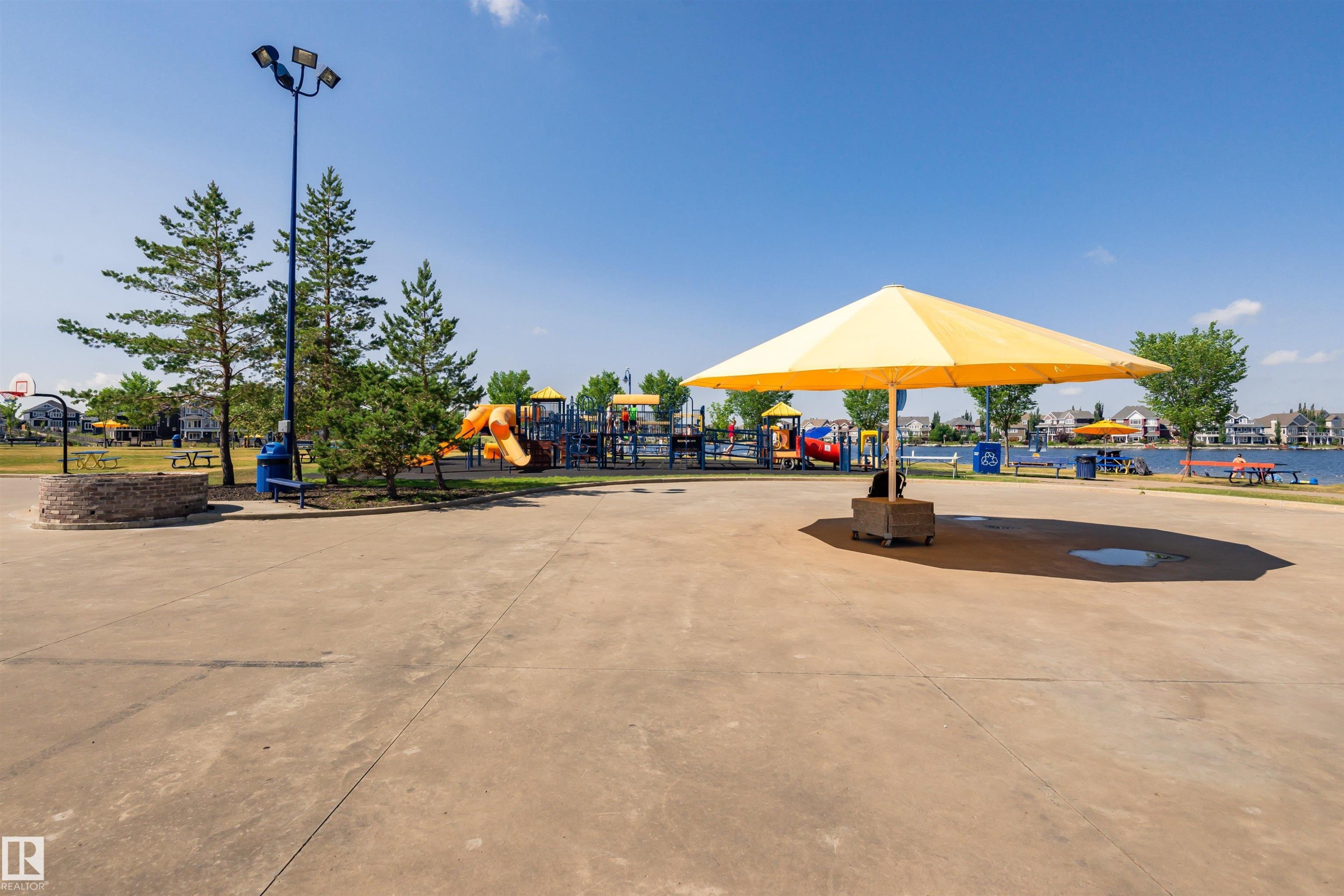 Spacious paved recreational area featuring a large yellow umbrella, mature evergreen trees, and a distant playground with slides - #66 8304 11 Av Sw, Edmonton, AB - Outdoor