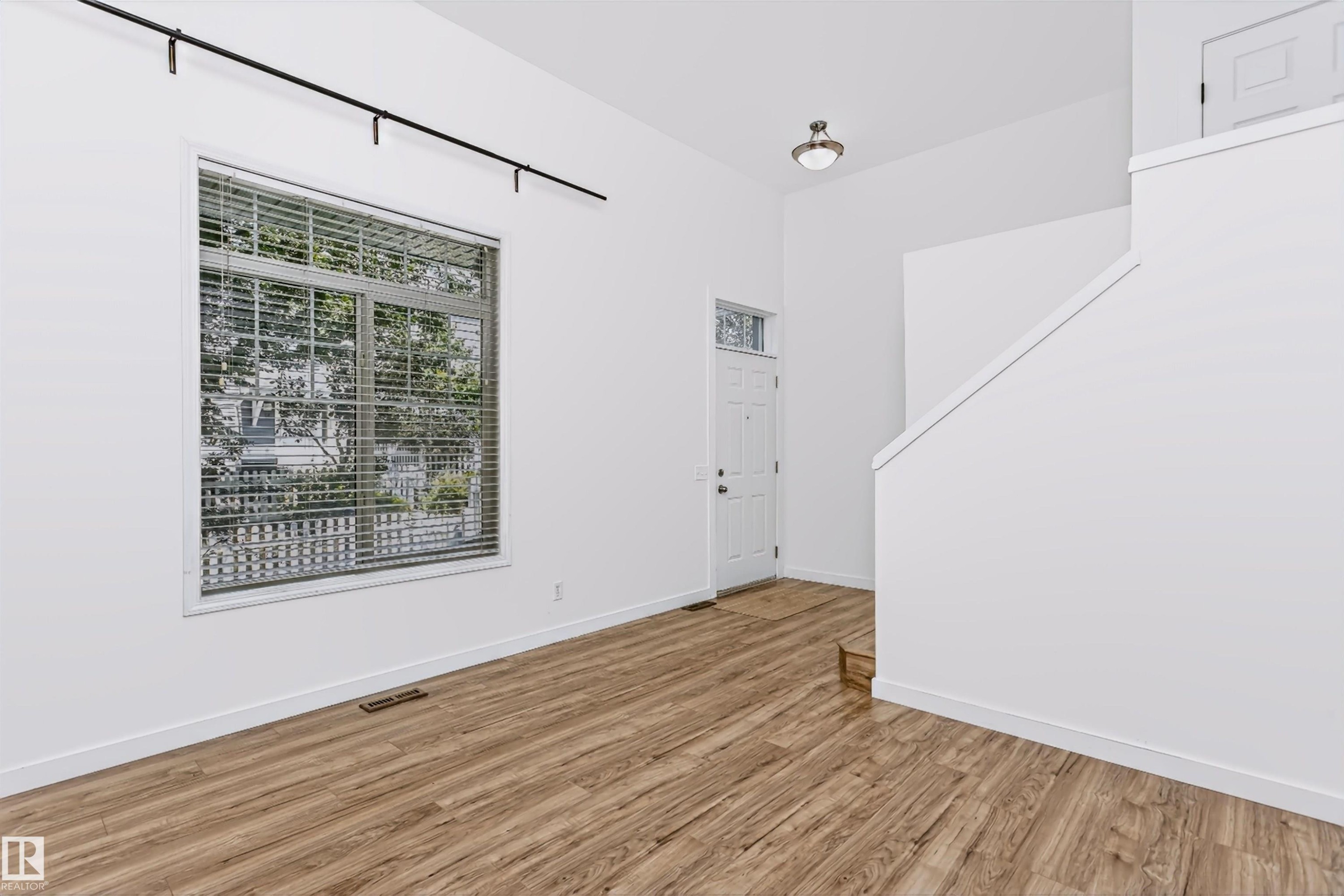 Open-concept living space featuring vaulted ceilings, wood-finish flooring, a contemporary window with blinds, a white entry door with a transom window, and a modern track lighting fixture - #66 8304 11 Av Sw, Edmonton, AB - Indoor Photo Showing Other Room