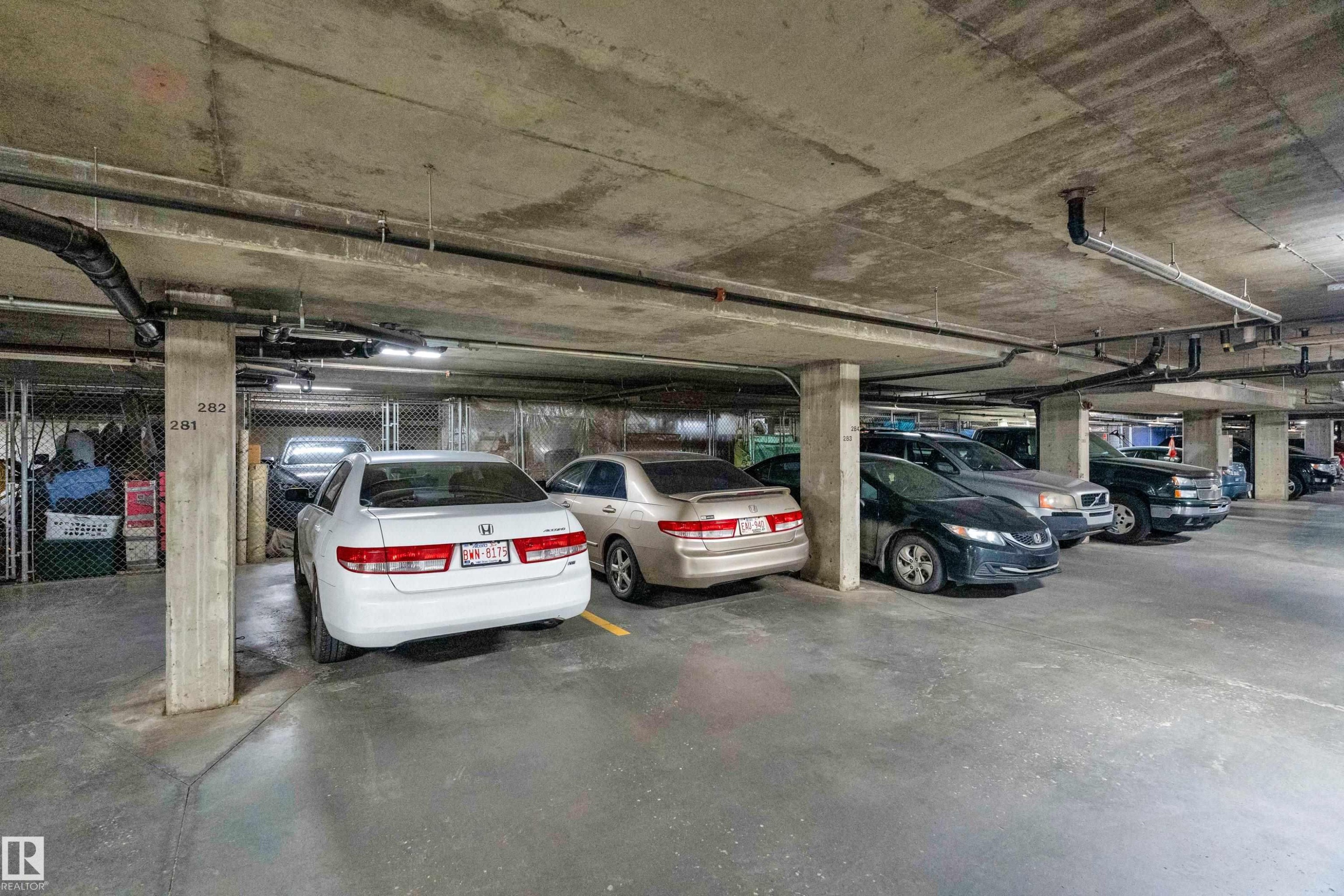 Underground parking garage featuring concrete pillars, exposed utility pipes, fluorescent lighting, and designated parking spaces - #136 16035 132 St Nw, Edmonton, AB - Indoor Photo Showing Garage