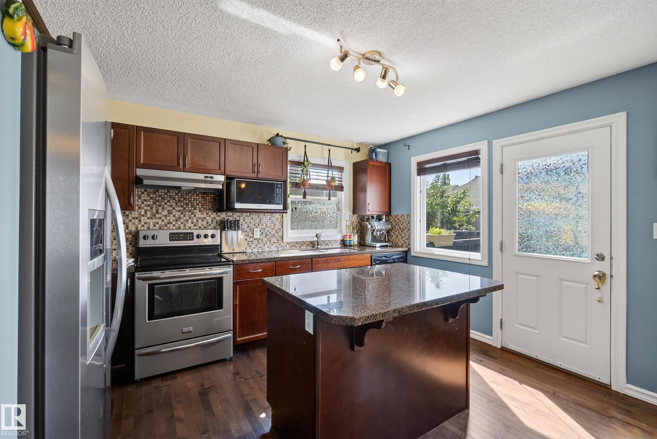 Kitchen featuring a central island with a stone-finish countertop, wood-finish cabinetry, stainless steel appliances, and a mosaic tile backsplash - 3003 20 Avenue Nw, Edmonton, AB - Indoor Photo Showing Kitchen With Double Sink