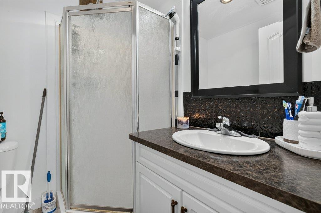 Bathroom featuring a glass-enclosed shower, white vanity cabinetry with dark hardware, a dark patterned backsplash, a countertop with an integrated sink, and a framed mirror - 12219 89 Street Nw, Edmonton, AB - Indoor Photo Showing Bathroom