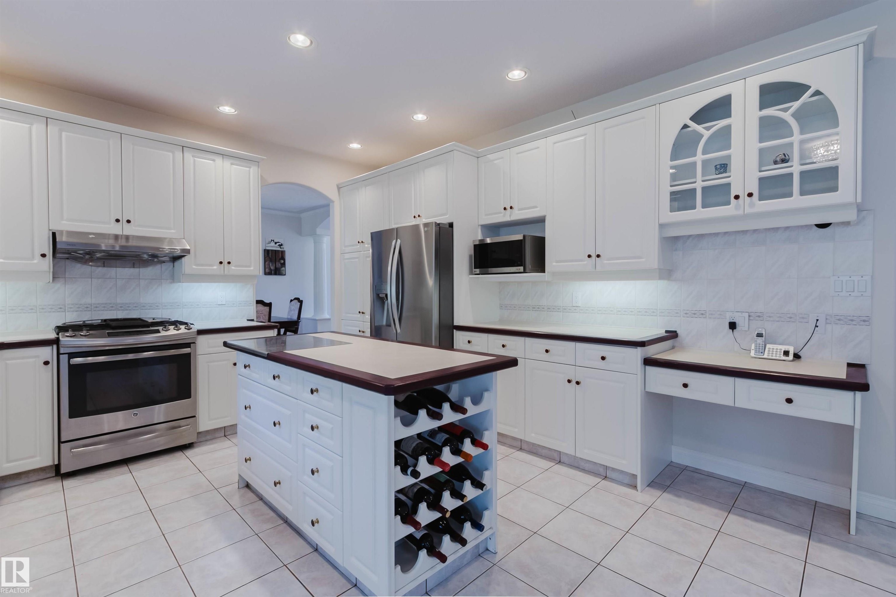 White kitchen cabinetry with stainless steel appliances, a central island featuring a built-in wine rack, light-toned tile flooring, and recessed lighting - 928 Heacock Road Nw, Edmonton, AB - Indoor Photo Showing Kitchen