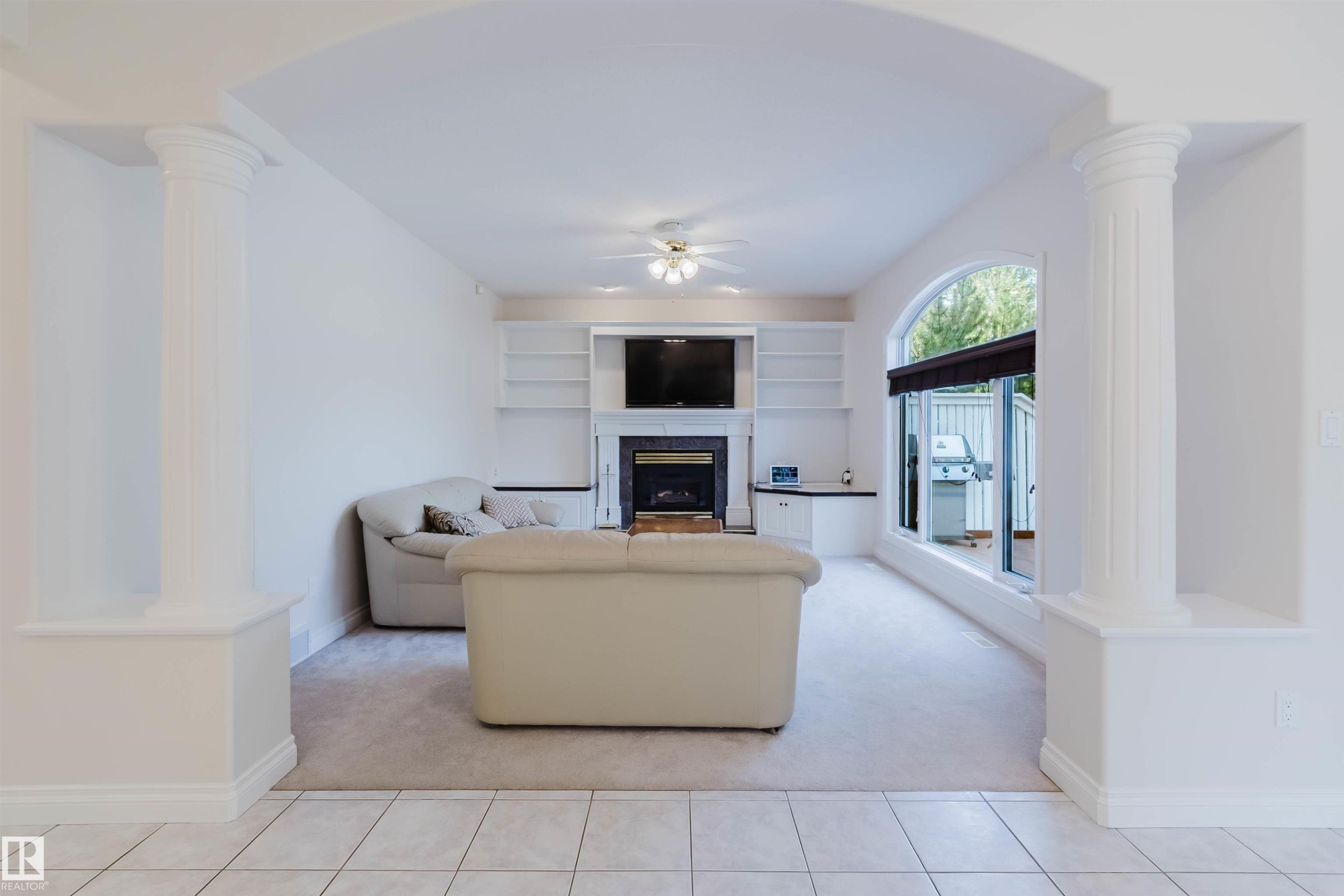 Living area featuring an arched entryway with decorative columns, a built-in fireplace with a dark surround, integrated shelving and cabinetry, and an arched window overlooking an outdoor patio - 928 Heacock Road Nw, Edmonton, AB - Indoor With Fireplace