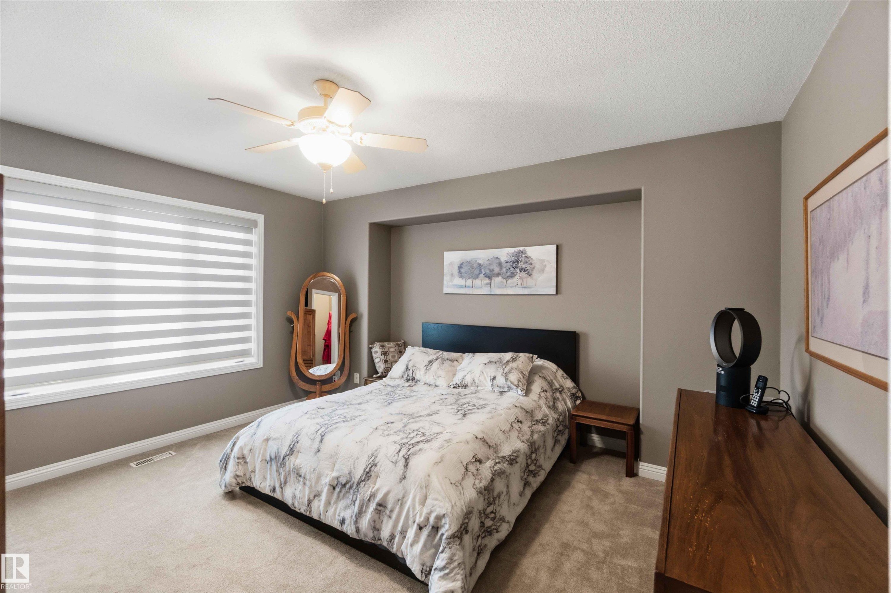 Carpeted bedroom featuring a large window with horizontal blinds, a ceiling fan with integrated lighting, and a recessed wall niche - 6320 166 Avenue Nw, Edmonton, AB - Indoor Photo Showing Bedroom
