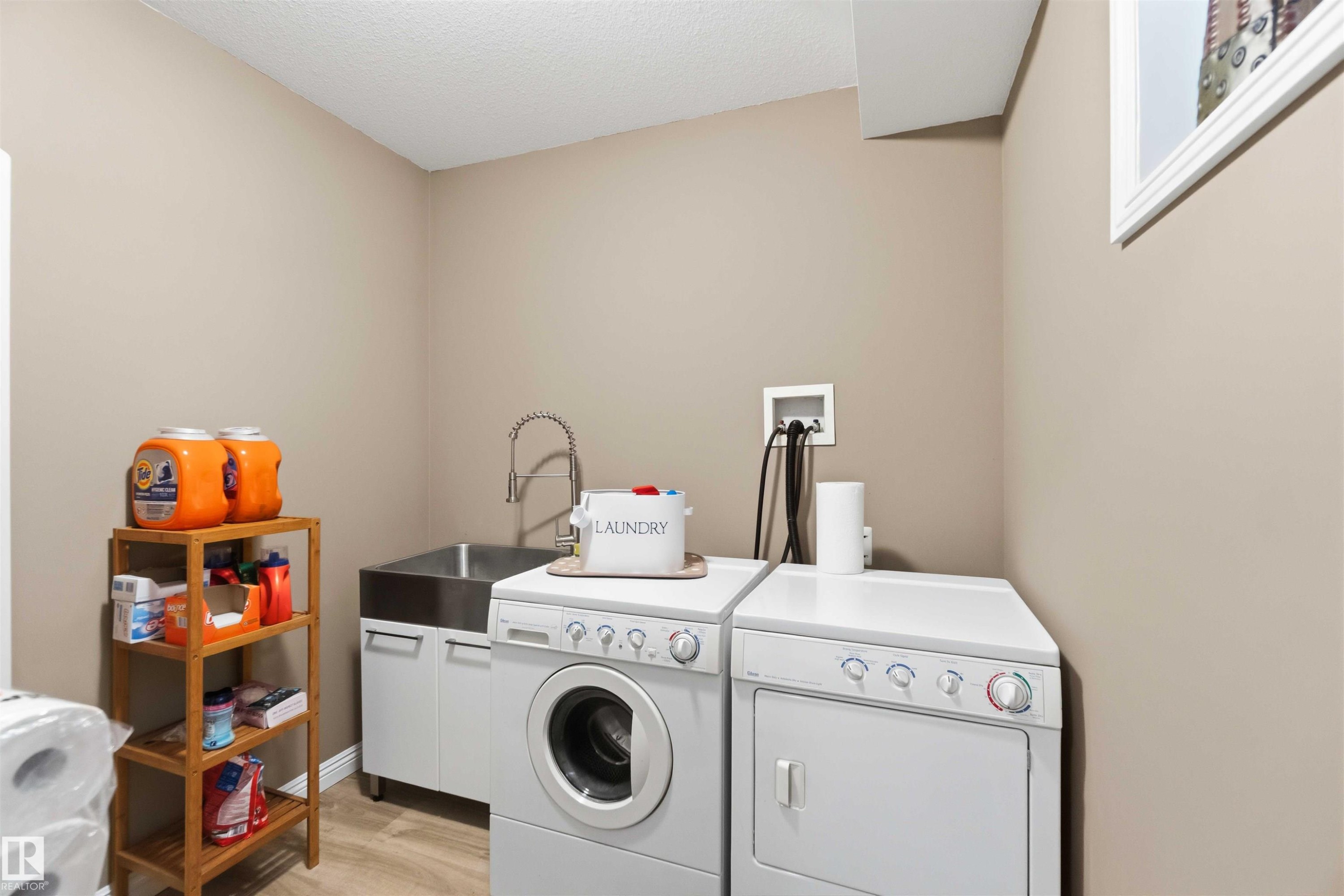 Dedicated laundry area featuring light-toned walls, a stainless steel sink with a gooseneck faucet, white cabinetry, and wood-finish flooring - 6320 166 Avenue Nw, Edmonton, AB - Indoor Photo Showing Laundry Room