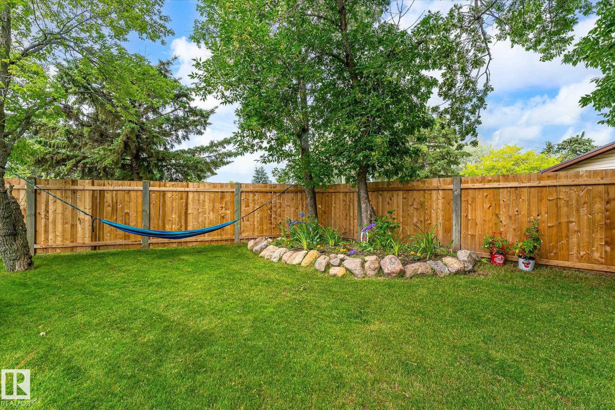 Spacious backyard featuring a wood fence, mature trees, green lawn, and a rock-lined garden bed with plantings - 4921 47 Avenue, Legal, AB - Outdoor With Backyard